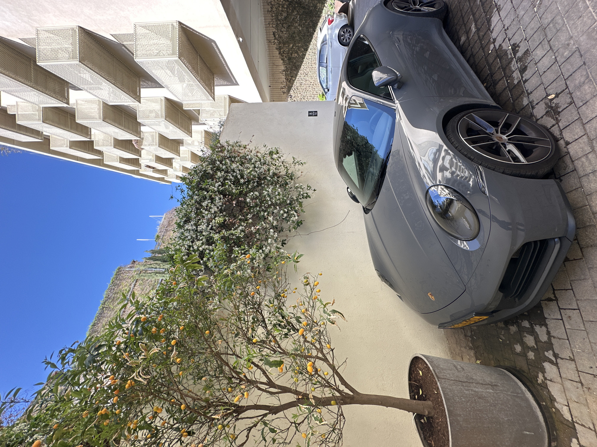 This media file captures an outdoor urban scene in Tel-Aviv, Israel, featuring a modern building, parked cars, and lush vegetation under a clear blue sky.
In the foreground, a sleek, dark gray Porsche sports car is parked on a sidewalk paved with light-colored rectangular tiles. The car's front section, including its distinctive headlights, part of the bumper, and a multi-spoke wheel, is prominently visible. A partially obscured license plate on the front bumper displays the letters "NJJ" followed by some numbers. Adjacent to the car, a kumquat tree, abundantly laden with small, yellow-orange fruits, grows in a large, textured metal planter.
To the left of the car and behind the kumquat tree, a vibrant bush with small white flowers cascades against a light-colored, plain wall. On this wall, a small black sign featuring a white wheelchair accessibility symbol is affixed, with the number "27" displayed directly below it.
The background is dominated by a modern building with a striking white facade. This facade is adorned with numerous horizontally extended, rectangular structures made of perforated material, creating intricate patterns and shadows. A bright, unblemished blue sky occupies the upper left portion of the image. Further back, a portion of another, lighter-colored car (possibly white or silver) is visible, parked behind the gray Porsche.
The scene is illuminated by bright daylight, casting crisp shadows and suggesting a sunny day. No people are visible, and the overall impression is one of a quiet, upscale urban environment.