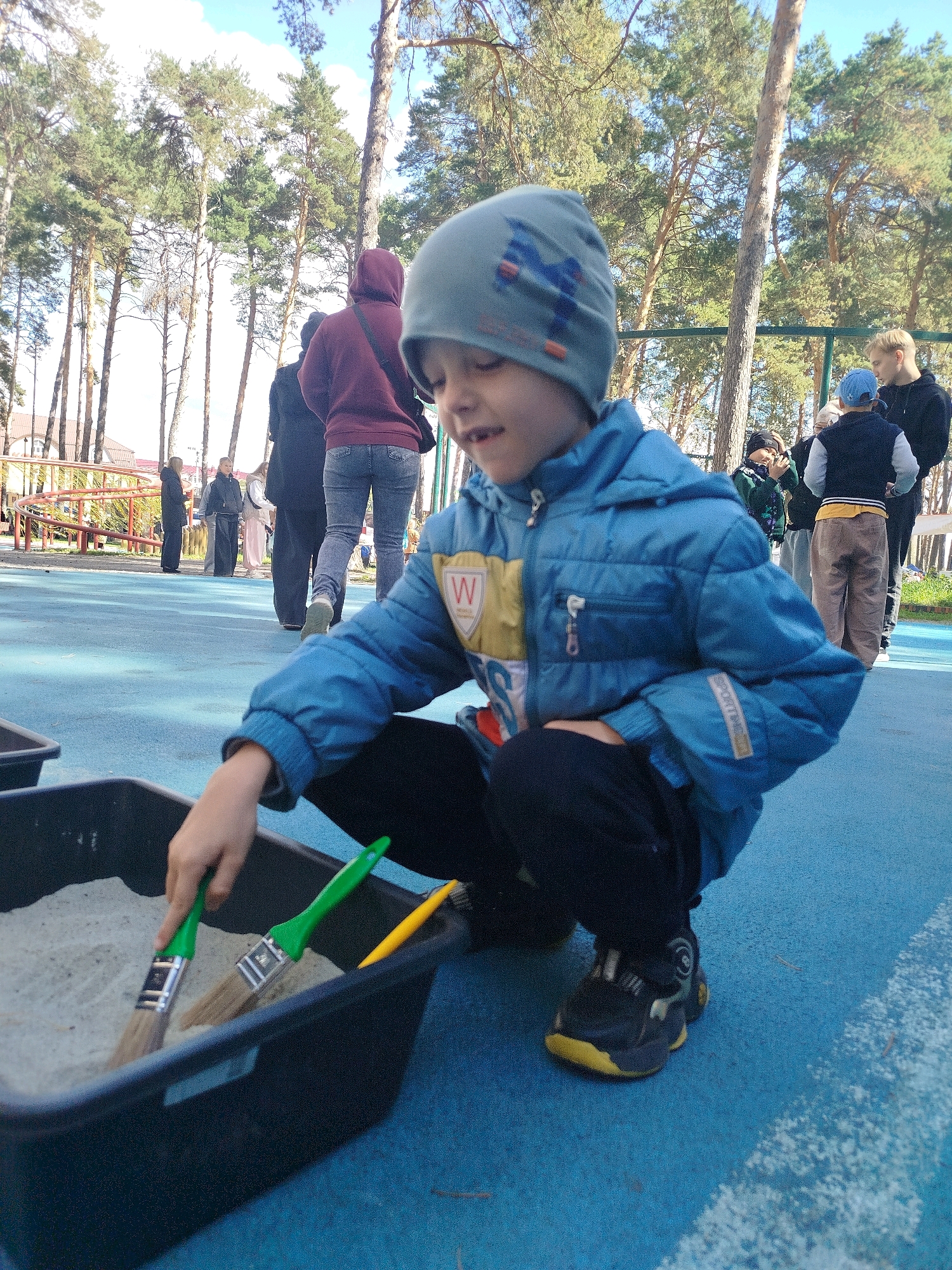 In Zavodoukovsk, Russia, a young boy, wearing a blue jacket and a gray beanie, is crouched down on a blue surface. He is actively engaged with a black plastic container filled with sand, using two green-handled brushes to sift through the material. A yellow object, possibly another tool, is also visible in the sand. The background features several individuals, some standing and observing, while others are in motion, suggesting a public outdoor area like a park or playground. Tall pine trees dominate the background, indicating a natural, possibly forested, environment. The lighting and clear sky suggest it is daytime.