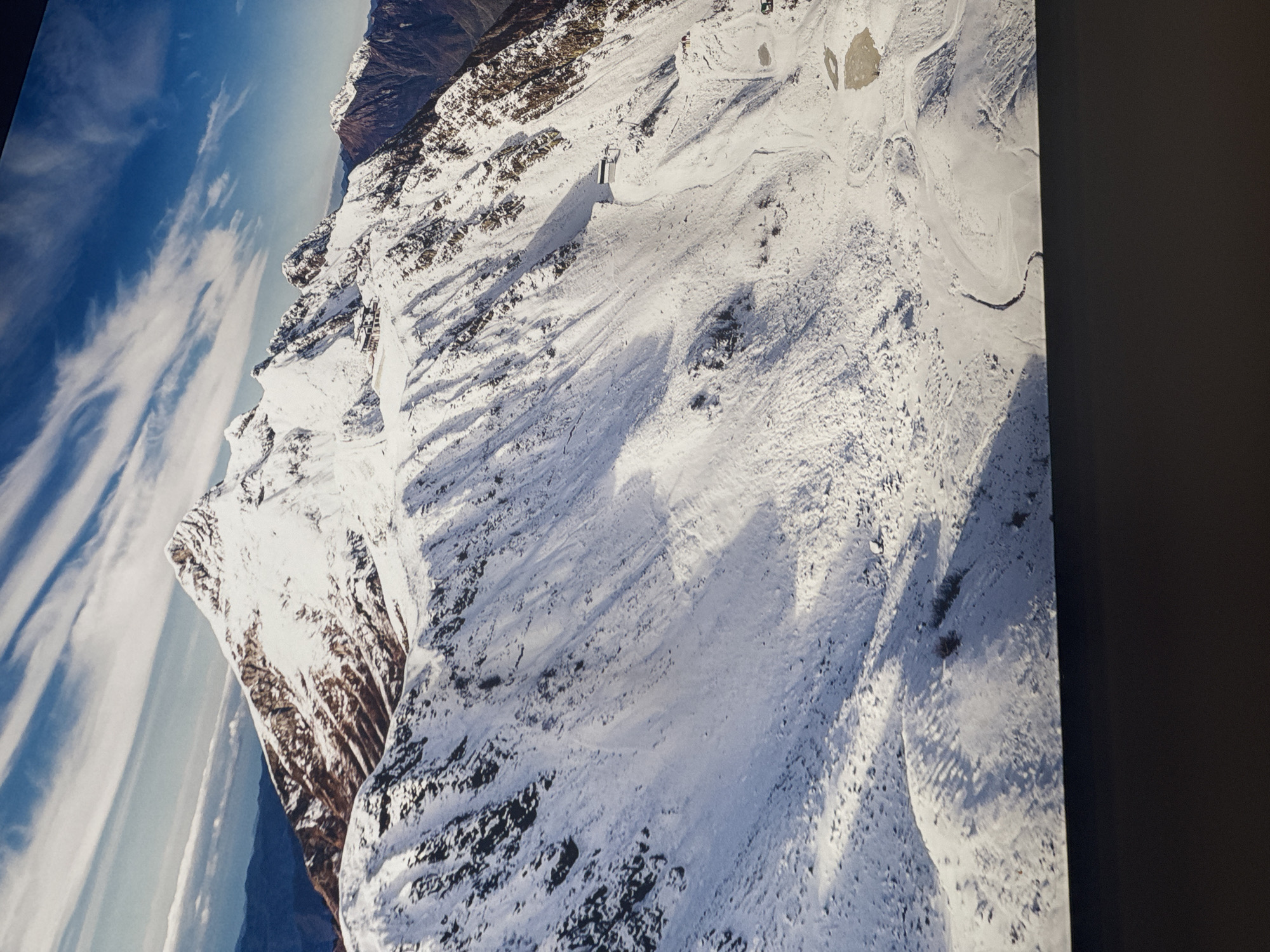 The image depicts a snow-covered mountain range under a blue sky with wispy clouds. The mountains have a mix of snow and dark rocky patches. A winding path or road is visible on the snow-covered terrain.
The location is likely within the Caucasus Mountains near Sochi, Russia, as the Rosa Khutor Alpine Resort is situated there and exhibits similar mountainous terrain and snow coverage.