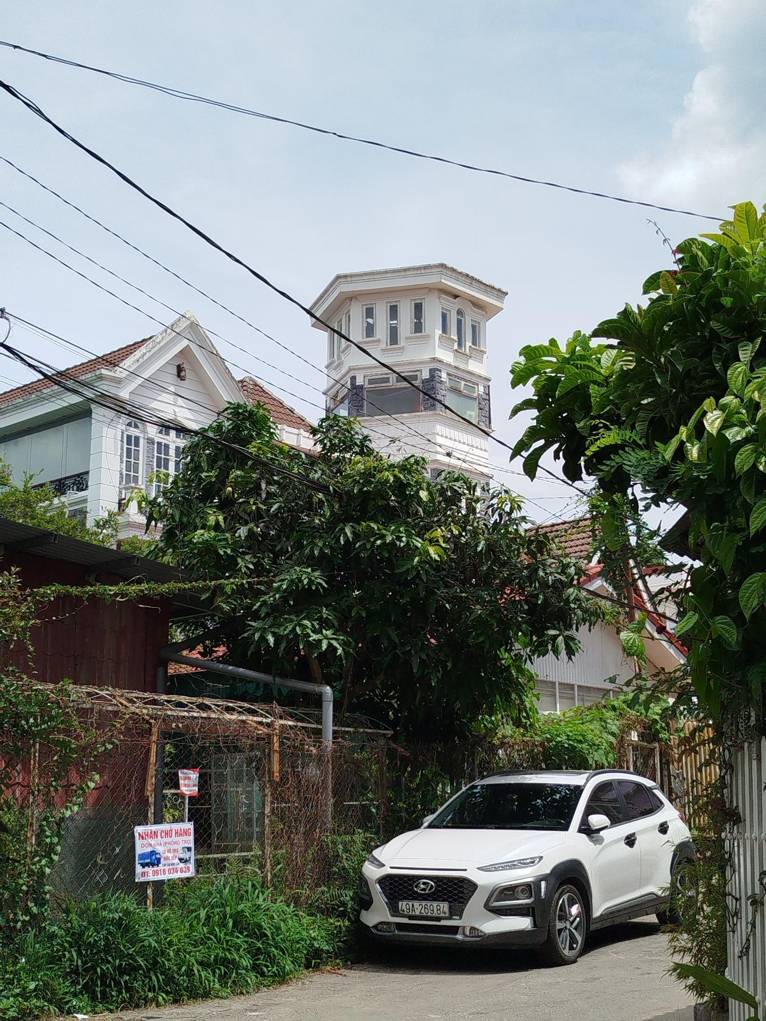 The image depicts a street scene in Da Lat, Vietnam, characterized by a mix of residential architecture, lush vegetation, and overhead power lines. In the foreground, a white Hyundai Kona SUV is parked on the right side of a narrow asphalt road. The car has the license plate "49A-269.84". To the left of the car, a rusty chain-link fence encloses a yard with overgrown green grass and some bushes. A white sign with blue and red lettering is attached to the fence, which appears to be an advertisement for delivery services.
Behind the fence, the landscape rises with dense green trees and the roofs of several houses. The most prominent structure is a multi-story building with a distinctive octagonal tower featuring multiple windows on the upper levels. Adjacent to it, a white house with a red-tiled roof and large arched windows is visible. The architecture suggests a blend of modern and traditional styles, common in Da Lat. Overhead, a complex network of black power lines crisscrosses the sky, a ubiquitous element in urban Vietnamese landscapes. The sky is overcast with scattered clouds. The overall impression is a residential street in a hilly, verdant city.