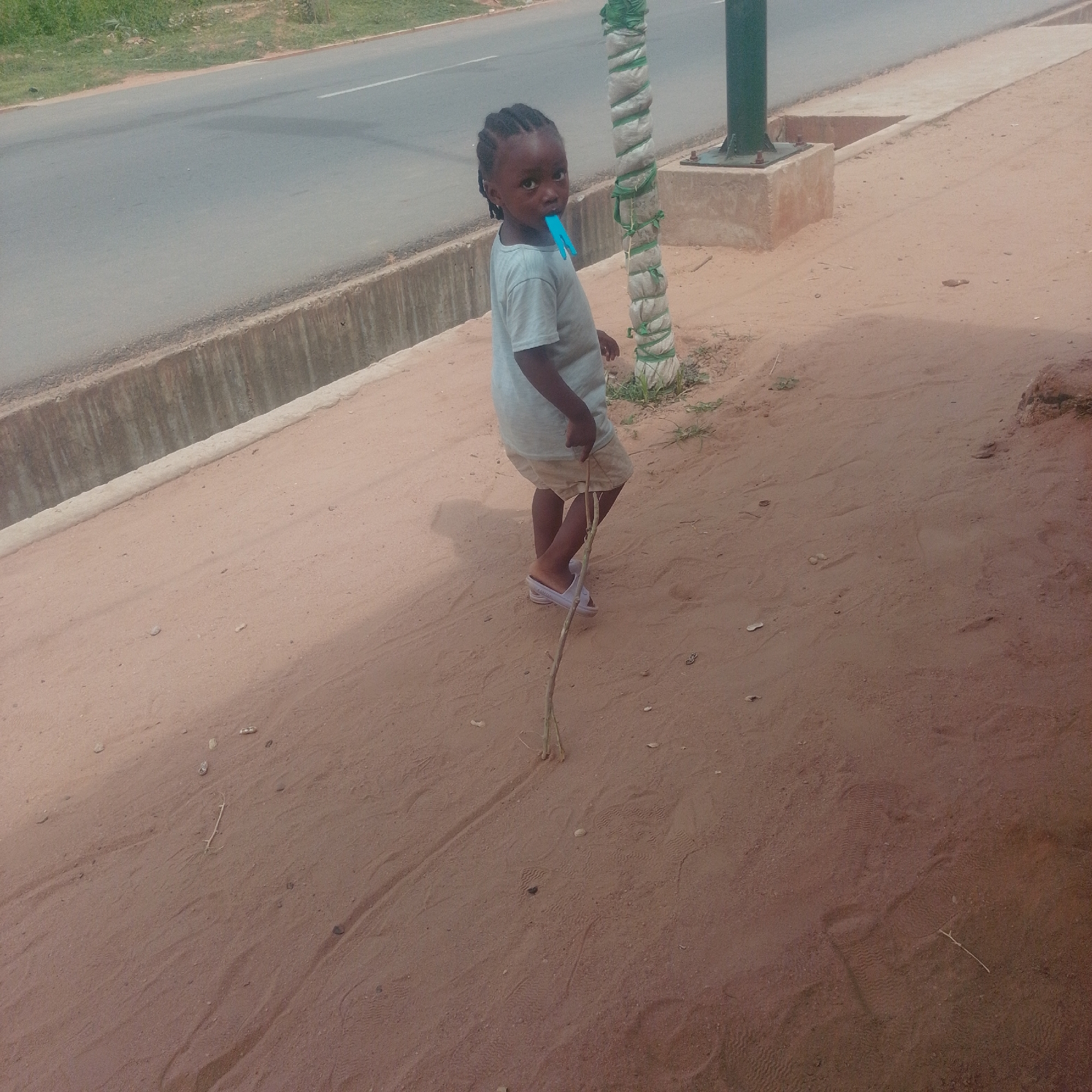 A young child with braided hair stands on a sandy path, holding a stick. The child is wearing a light-colored t-shirt and shorts, and light-colored sandals. A blue object, possibly a clothespin, is in the child's mouth. The child is looking towards the camera. Behind the child is a pole wrapped in white and green material, with some grass growing at its base. To the left of the child is a concrete structure and a paved road with a white line, bordered by a concrete drainage channel and green grass. The ground is a reddish-brown sand with some scattered debris and footprints. The lighting suggests it is daytime, with the sun casting shadows. The overall scene appears to be an outdoor setting in a possibly rural or suburban area, given the presence of the road, drainage, and sandy ground. The child's expression is neutral. There is no visible text in the image.