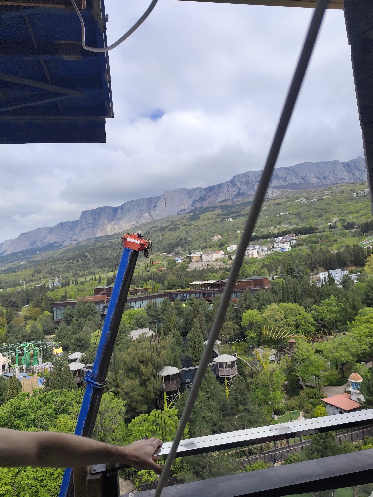 The image depicts a view from an elevated platform in Симеизский поселковый совет, Ukraine, overlooking a mountainous landscape and a wooded area containing buildings and structures, including what appears to be a miniature golf course and a small amusement park. A construction crane with a blue boom and red head is partially visible in the lower left. A hand is holding onto the railing. The sky is overcast.