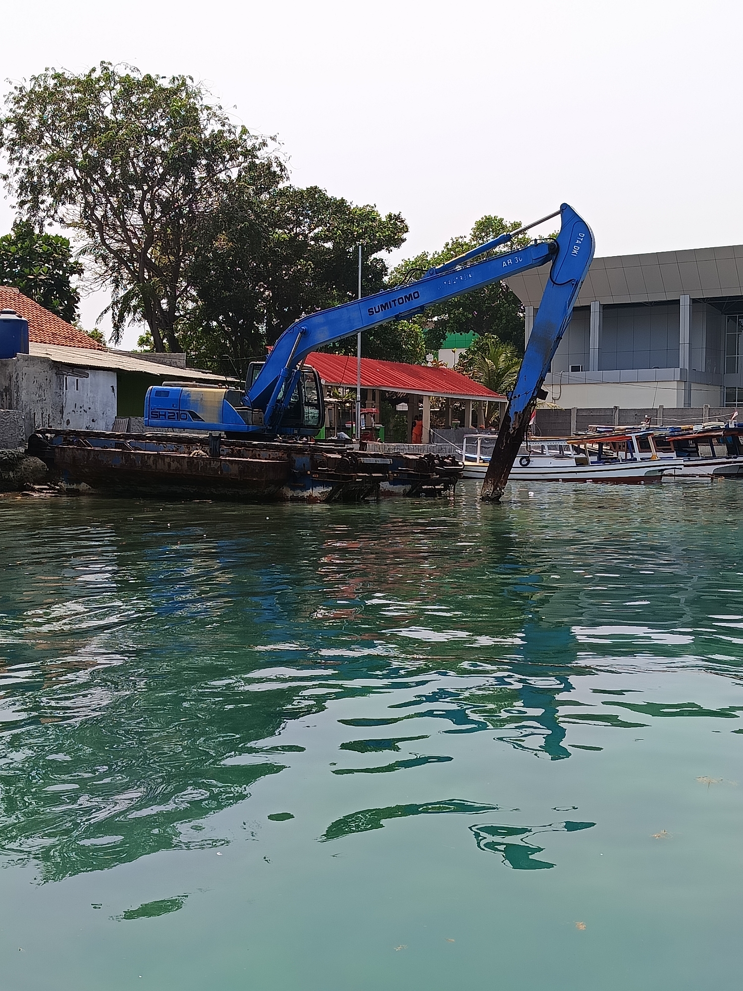 A blue excavator is mounted on a barge in the water, near a harbor. The excavator is actively digging or working on something underwater. Several boats are docked in the background, along with buildings and trees. The water is calm and reflects the sky and the surrounding structures. The scene appears to be in an urban or semi-urban coastal area.