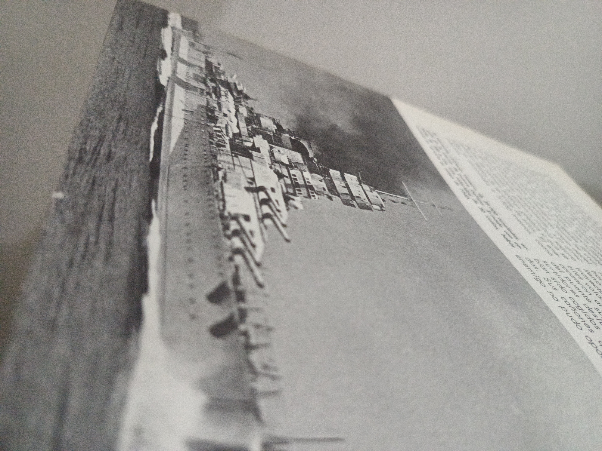 A close-up, black and white image of a book laid open. The left page displays a picture of a large ship at sea, while the right page contains text in a language that appears to be Spanish. The ship's structure is complex, with many protruding elements. The text on the right mentions words like "enemigo" (enemy), "puente" (bridge), and "cañones" (cannons), suggesting a war-related theme. The image is likely a photograph of a page in a book, possibly about naval history or warfare. The location, Los Robles, Venezuela, is not discernible from the image.
