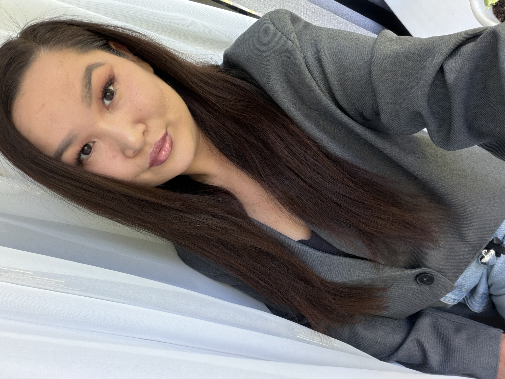A selfie depicts an Asian woman with long, straight, brown hair, wearing a grey blazer. The background consists of white, translucent fabric. The setting is Unknown, Unknown.
