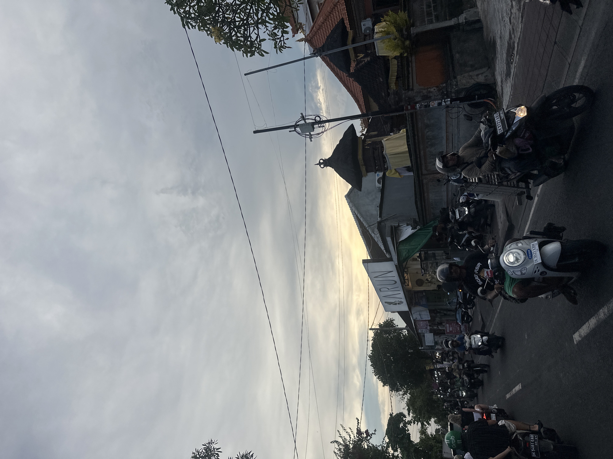 The image depicts a street scene in North Kuta, Indonesia. The view is oriented at an angle, showing the sky and the side of a road congested with parked and moving motorbikes. A building with a Balinese architectural style is visible on the right. Overhead electrical wires stretch across the sky. A sign for "ARUN" is seen on a building, and various businesses line the street. The sky is overcast. Several people are riding motorbikes; close-up license plates read "DK 5401 GAB," "DK 3521 KBM," and "DK 3723 ABW." One rider is wearing a helmet and a shirt with "SURF ASIA CHAOS" printed on it.