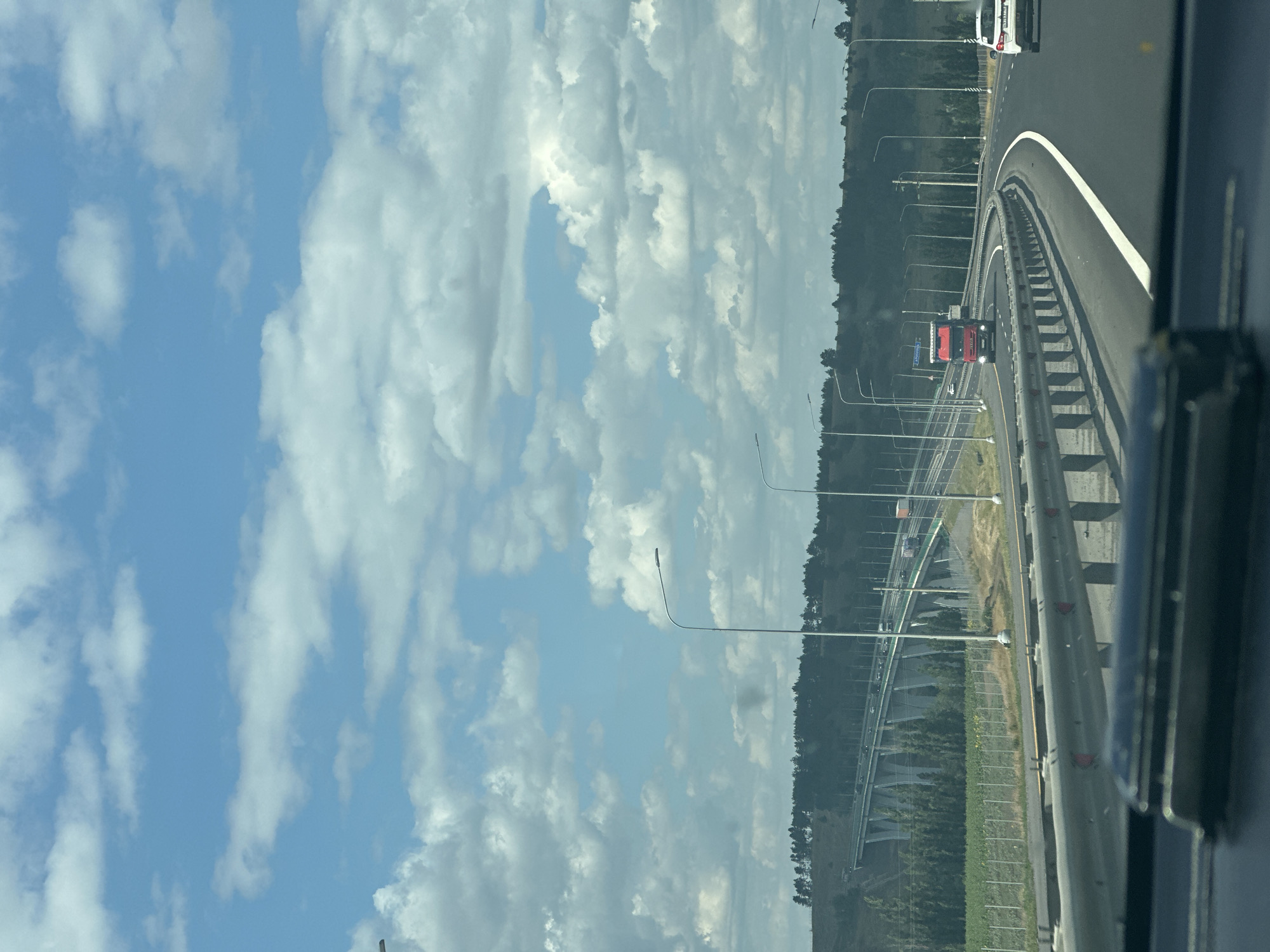The image captures a highway scene with a bright blue sky filled with fluffy white clouds. A multi-lane highway curves to the right, bordered by a metal guardrail. On the left side of the highway, a large elevated bridge with a series of angled concrete supports is visible, with vehicles traveling on it. A dense line of dark green trees runs along the crest of a hill behind the bridge. On the highway itself, a red truck is driving in the left lane, and a white car is in the right lane. Streetlights line the side of the road. The foreground shows the interior of a vehicle, with a dark dashboard and a shiny object on it. The scene appears to be from a moving vehicle, suggesting it was taken while driving. Based on available information, this highway is likely part of the R-217, also known as the "Kavkaz" federal highway, near the settlement of Tarasovskii in Russia.