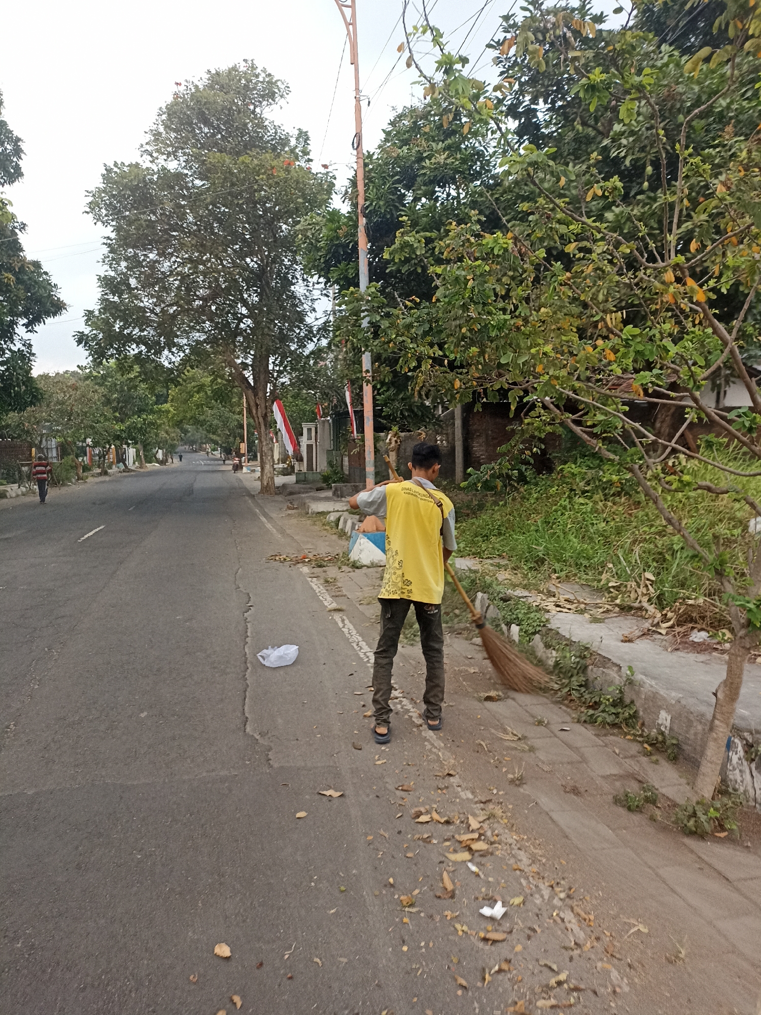 In Mojopanggung, Indonesia, a person in a yellow uniform is sweeping the side of a paved road with a broom. The individual is facing away from the camera, wearing dark pants and sandals, and is actively engaged in sweeping dry leaves and debris from the roadside onto the street. The environment features a road with a sidewalk to the right, bordered by vegetation and trees. In the background, there is a street with other people walking and a few vehicles. Red and white Indonesian flags are visible on poles along the sidewalk in the mid-ground. The sky is overcast, suggesting a cloudy day.