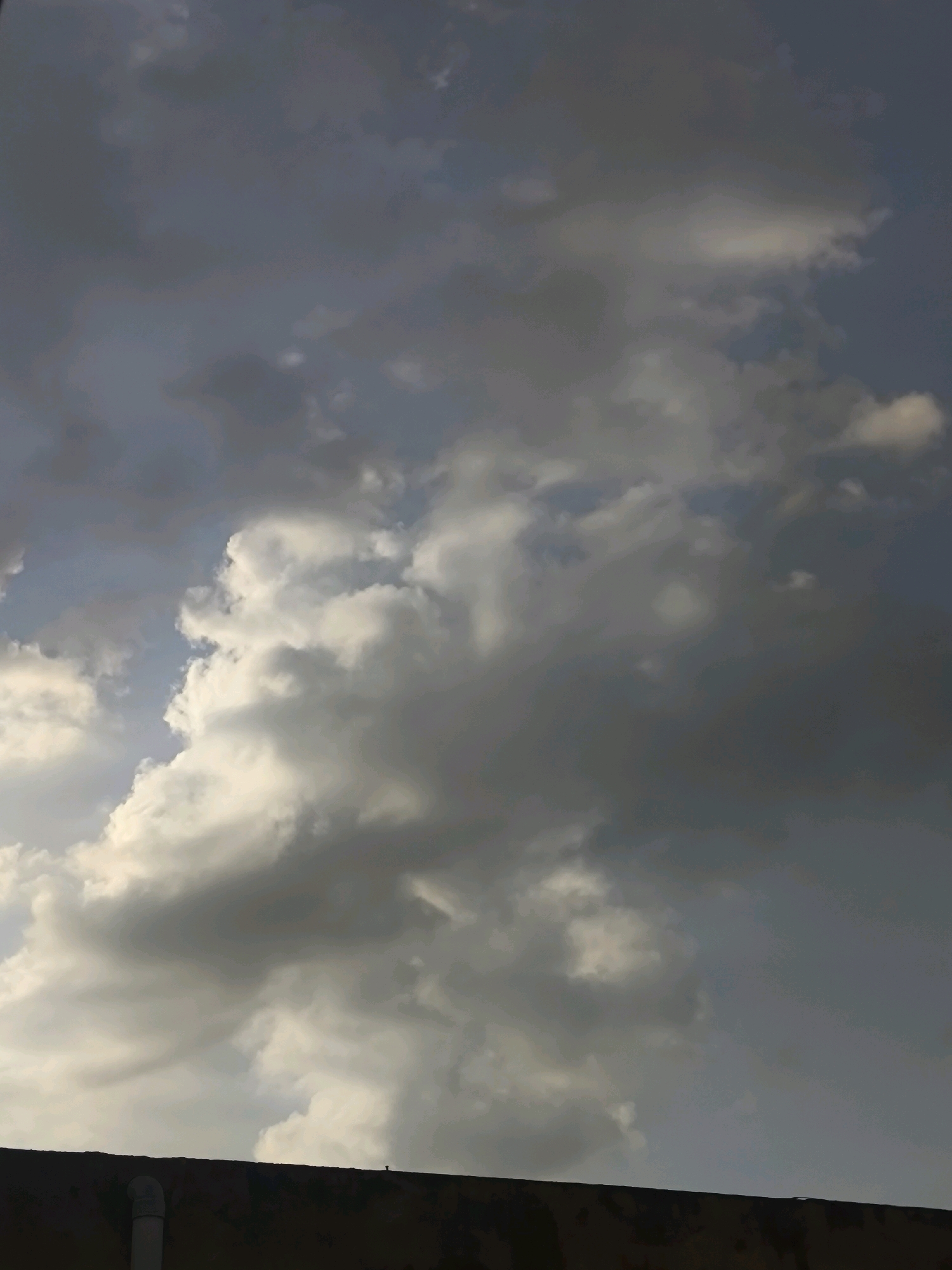 The image shows a cloudy sky above a dark, textured rooftop in Chhajpur Kalan, India. The clouds are a mix of white and dark gray, with some areas illuminated by sunlight and others casting shadows. A white pipe is visible on the left side of the rooftop, extending upwards. The overall impression is of an overcast sky with dramatic cloud formations.