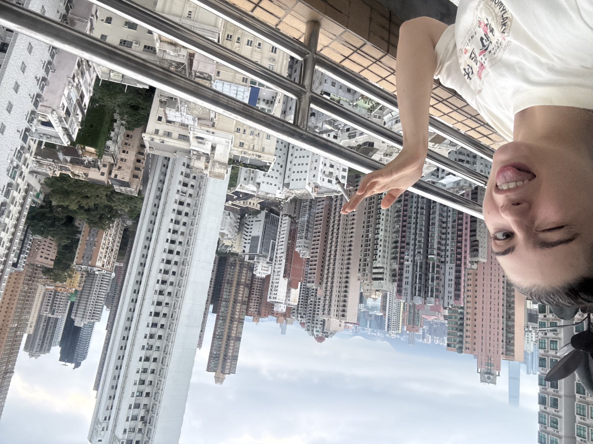 The image shows a woman's face from an upside-down perspective. She is wearing a white t-shirt and sticking her tongue out while winking. Behind her is a view of high-rise buildings, characteristic of a dense urban environment. The architecture and skyline suggest the location is Hong Kong, China. The foreground includes a metal railing, indicating the woman is likely on a balcony or rooftop. The sky is overcast and the buildings are of various heights, colors, and architectural styles.