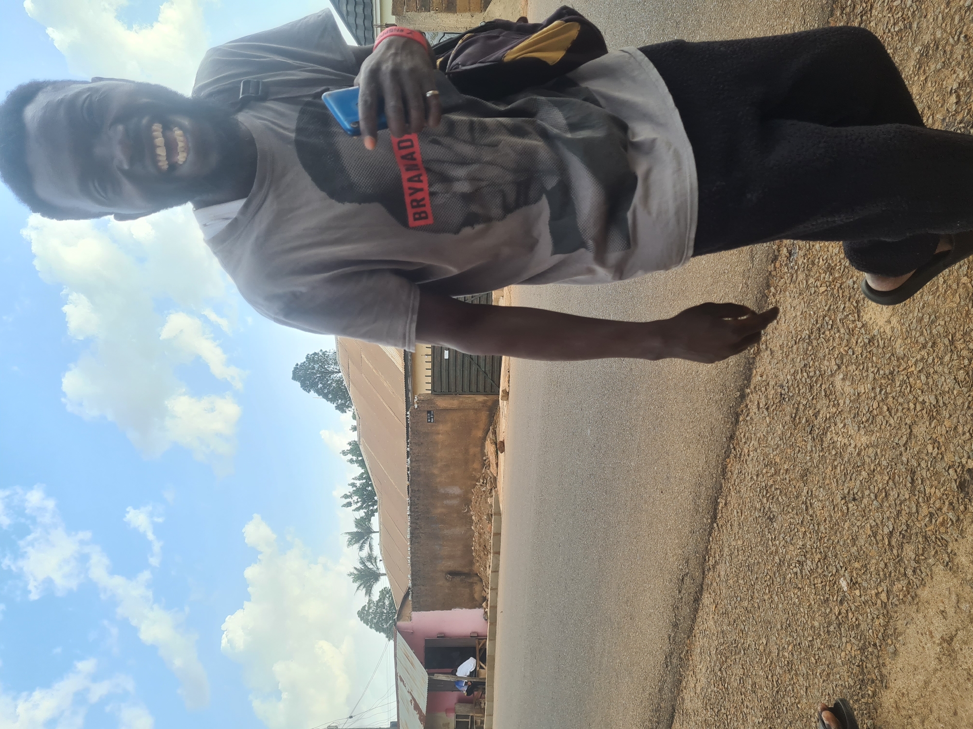 A man stands in the street, wearing a gray t-shirt with the word "BRYANAD" printed on it, along with black pants and sandals. He holds a blue cell phone in his left hand. A building runs lengthwise behind the man, situated on a paved road. Across the road is a patch of gravelly earth. The sky shows scattered clouds. The location is Jos, Nigeria.