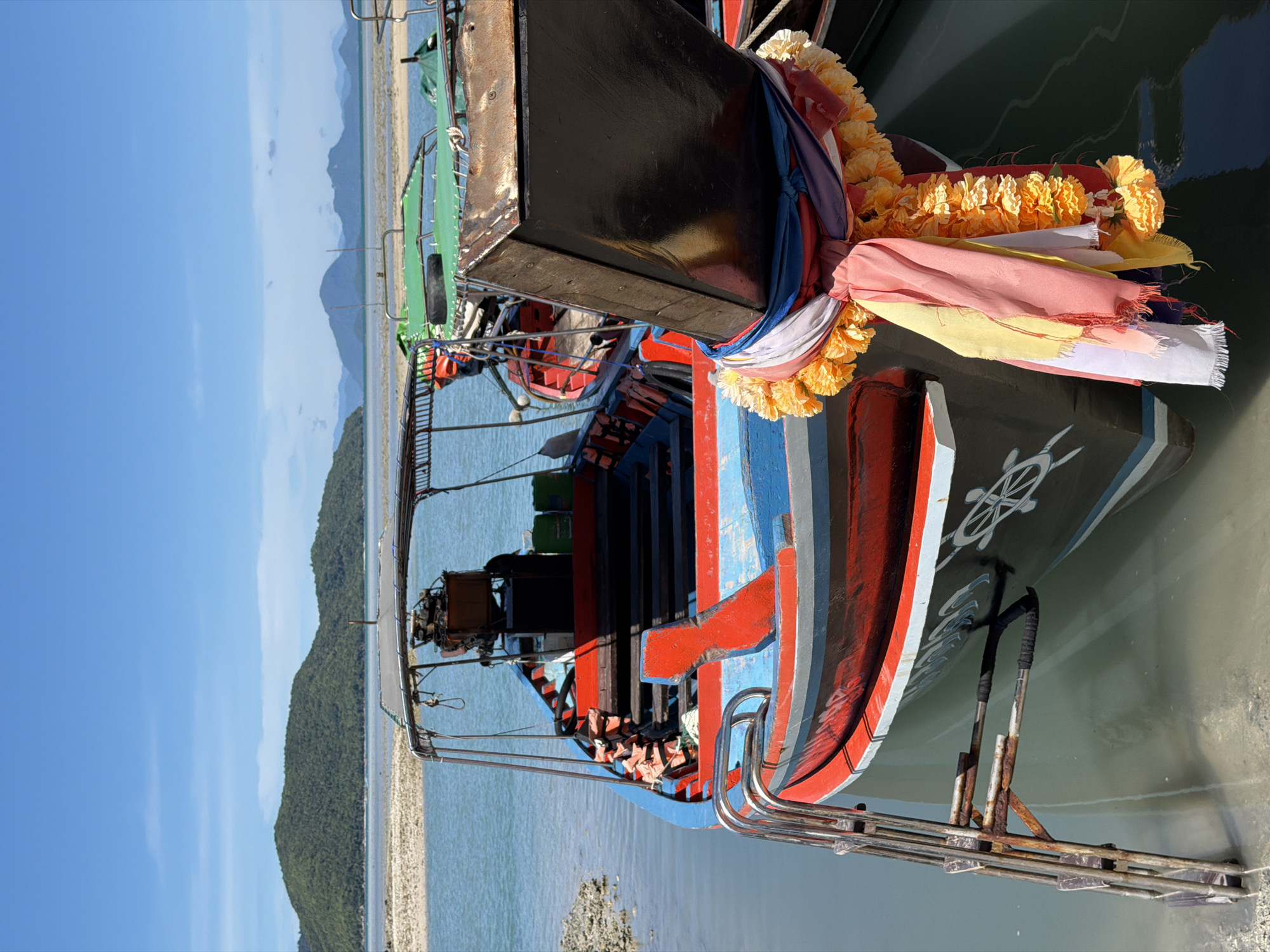 This media file depicts a brightly colored, traditional longtail boat docked in shallow, clear water. The boat is adorned with a garland of yellow marigolds and flowing fabric strips in various pastel colors, suggesting a festive occasion or religious offering. The hull of the boat is painted in bold colors of blue and red, with a white steering wheel emblem visible on its side. Several life vests are neatly stacked inside the open passenger area.
In the background, a sandy beach lines a lush, green, tree-covered hillside that slopes gently towards the water. The sky is a clear, bright blue with a few wispy clouds, indicating a sunny day. The water is calm and reflects the blue of the sky. The scene suggests a tropical location, consistent with the provided location context of Baan Thong Krut, Thailand. The lighting and clear skies suggest it is daytime. There is no explicit activity or event taking place, beyond the boat being docked. The overall impression is one of tranquility and vibrant local culture.