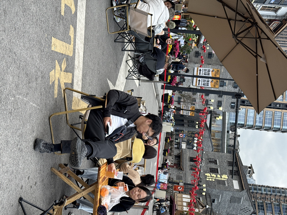 The scene depicts a vibrant outdoor dining and pedestrian area within Yuzhong District, China, under an overcast sky during daytime. The ground is asphalt, with yellow painted Chinese characters visible, suggesting a former or current roadway adapted for pedestrian use.
In the immediate foreground, a male subject is seated in a light-colored portable folding chair, facing left in profile. He wears a black long-sleeved jacket over a white t-shirt, dark trousers, dark socks, and black athletic shoes. A dark messenger bag with an orange accent is slung across his chest. He holds a dark smartphone in his left hand, gazing downwards at the screen, with his right foot resting on his left knee. To his right, a female subject is seated at a small, light-colored wooden folding table, also in a portable folding chair. She wears a dark jacket and holds a clear plastic cup containing a light-colored beverage in her right hand. Her gaze is directed downwards, possibly at a device obscured by a clear plastic bag on the table that contains several orange fruits.
In the mid-ground to the left, beneath a large, beige cantilevered umbrella, another group of individuals is seated at similar portable tables and chairs. This group includes at least six people engaged in various activities, such as looking at phones or conversing. One male subject with a green beanie and a white jacket is visible, smiling towards the camera.
The background features a mix of architectural styles. Several multi-story buildings constructed with grey stone-like facades and traditional sloped roofs are visible, interspersed with taller, modern high-rise buildings featuring glass and steel construction. Red decorative lanterns are strung across the street or pedestrian thoroughfare, adding a festive element. Streetlights and various signs with Chinese characters are present on poles; one sign clearly displays "新店开业" (New store opening) and "桂林台球" (Guilin Billiards). A yellow barrier cordons off an area to the left, behind which flower beds containing pink blooms are visible. Numerous other pedestrians are visible moving through the area in the background.