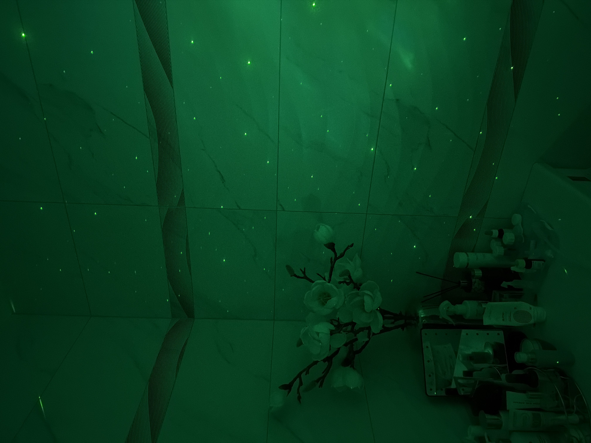 The image depicts a bathroom interior under green lighting. The walls are tiled with marble-patterned tiles, some featuring vertical striped accents. A bouquet of white flowers lies on the tiled floor near the wall. To the right, a cluttered shelf contains various toiletries and cosmetic products. The scene is observed under a consistent, saturated green illumination with what seems to be fiber optic lighting. The location within Votkinsk, Russia cannot be determined based on the provided image.