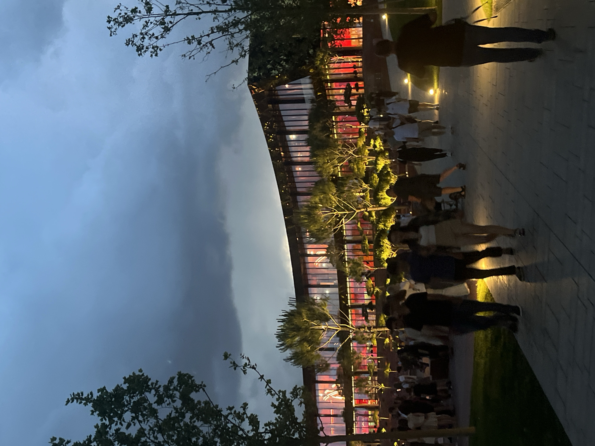 The image shows a group of people walking along a paved pathway in front of a modern building with large windows. The building is illuminated from within with a reddish glow, and the exterior is lined with trees and decorative lighting. The sky is dark and appears to be cloudy. The scene suggests an evening gathering or event at a public space, possibly a park or an entertainment venue. Based on the location provided, this scene is likely at Park Galitsky in Krasnodar, Russia, which is known for its contemporary architecture and well-maintained green spaces.