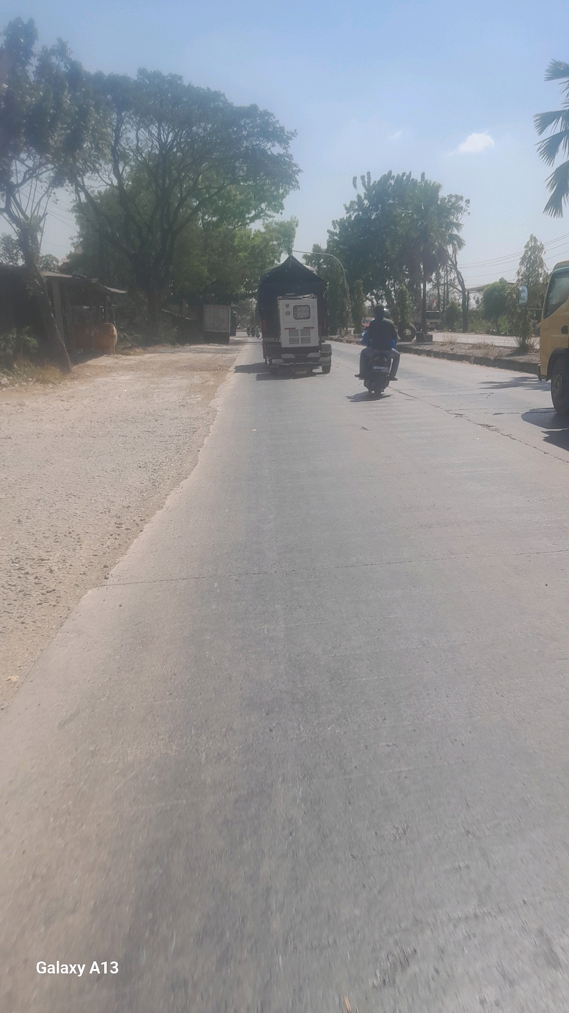 The image depicts a street scene in Tanjunggemuk, Indonesia. A light-colored, paved road occupies the majority of the frame, with a dusty, unpaved shoulder on the left. A white truck with a dark tarp covering its cargo is traveling away from the viewer in the right lane. To the right of the truck, a person on a motorcycle is also moving away from the viewer, wearing a dark shirt. In the background, numerous trees line the roadside, with some structures visible behind them. The sky is a clear, pale blue with a few wispy clouds. The lower left corner of the image displays the text "Galaxy A13".