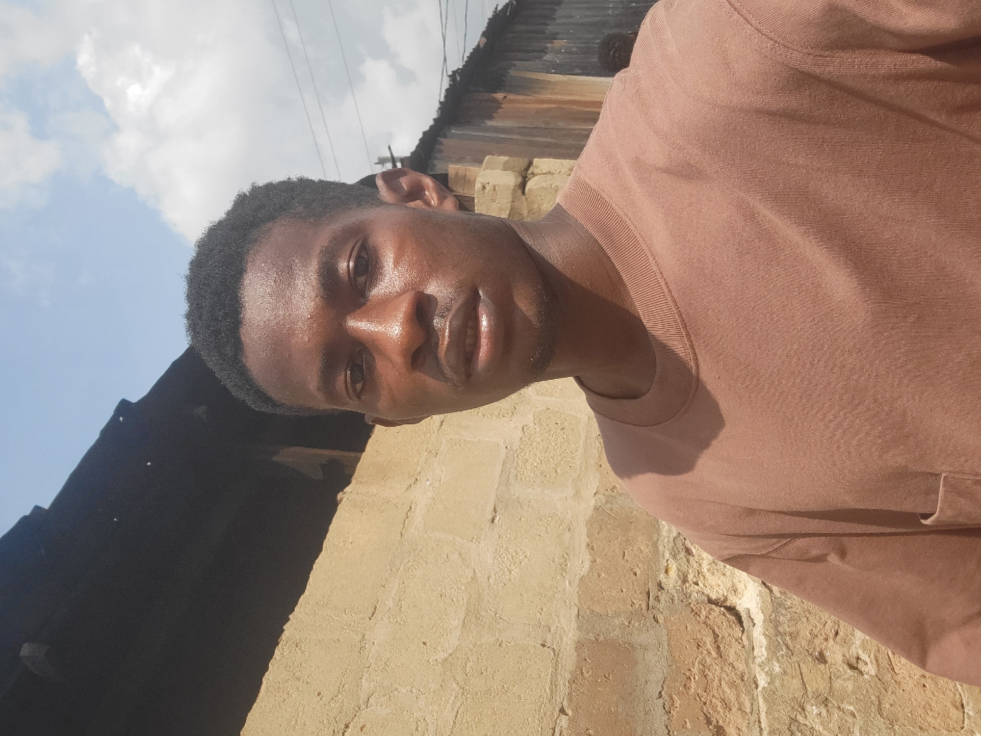 A young Black man, appearing to be in his late teens or early twenties, takes a selfie in an outdoor, urban environment. He has short, dark, curly hair and is wearing a light brown crewneck t-shirt with a pocket on the left chest. His skin is glistening with sweat. He is standing in front of a light brown brick wall, which appears to be part of a building. To the right of the brick wall, there is a darker structure with corrugated metal siding. Above, the sky is visible with scattered white clouds. The lighting suggests it is daytime, likely during hot weather, given the man's perspiration. The background, with its weathered construction materials and the glimpse of a power line, suggests a residential or informal settlement area, consistent with locations in Egbagu, Nigeria.
