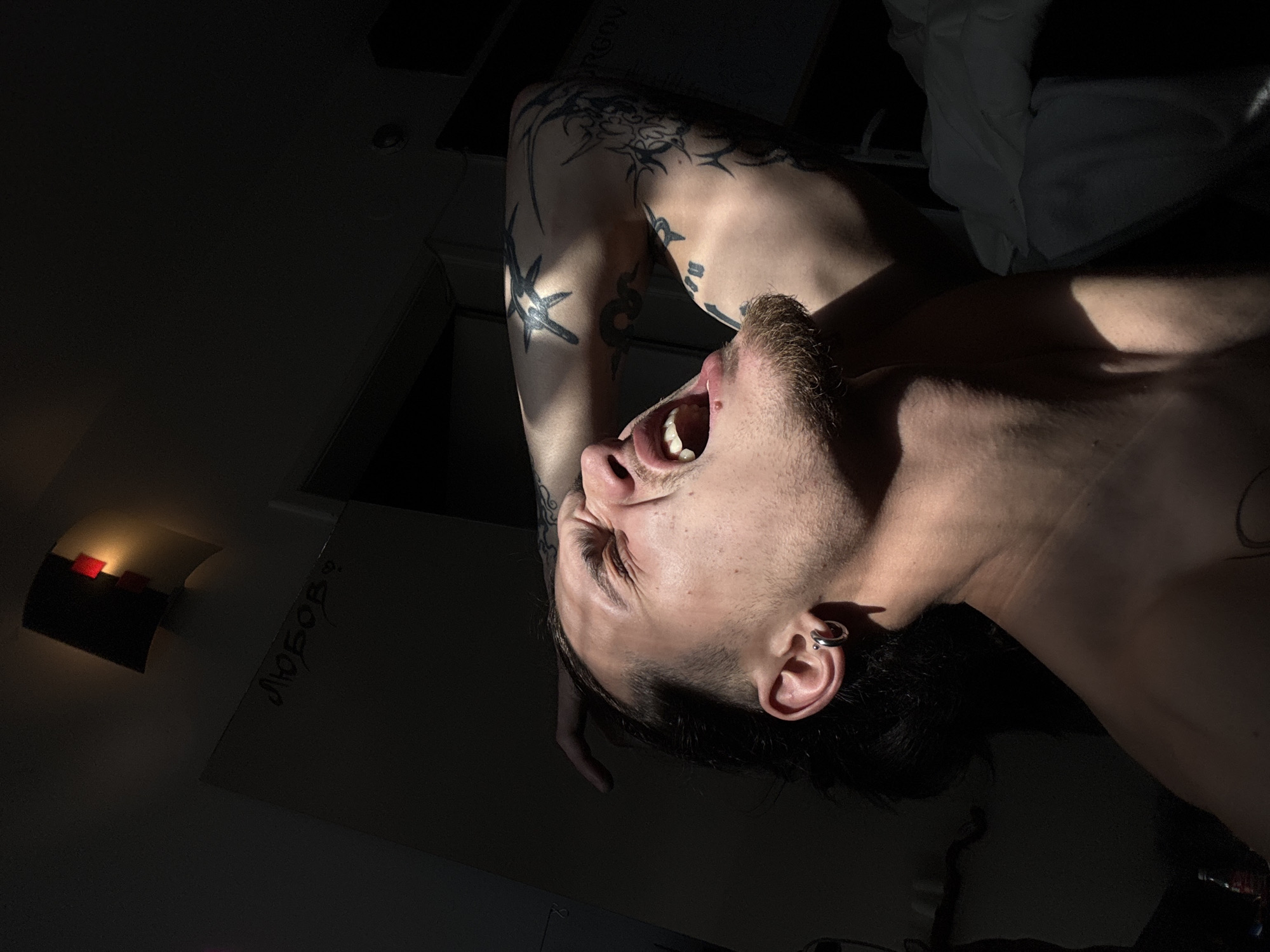 A low-angle, rotated shot captures a shirtless man with a goatee and tattoos on his left arm, his head tilted back, mouth open in a yell. The environment is dark, illuminated by a strong directional light source. A wall-mounted light fixture and a note written on the wall are barely visible in the background. The location is unclear. It is impossible to determine if the photo was taken in Sofia, Bulgaria, without further information.