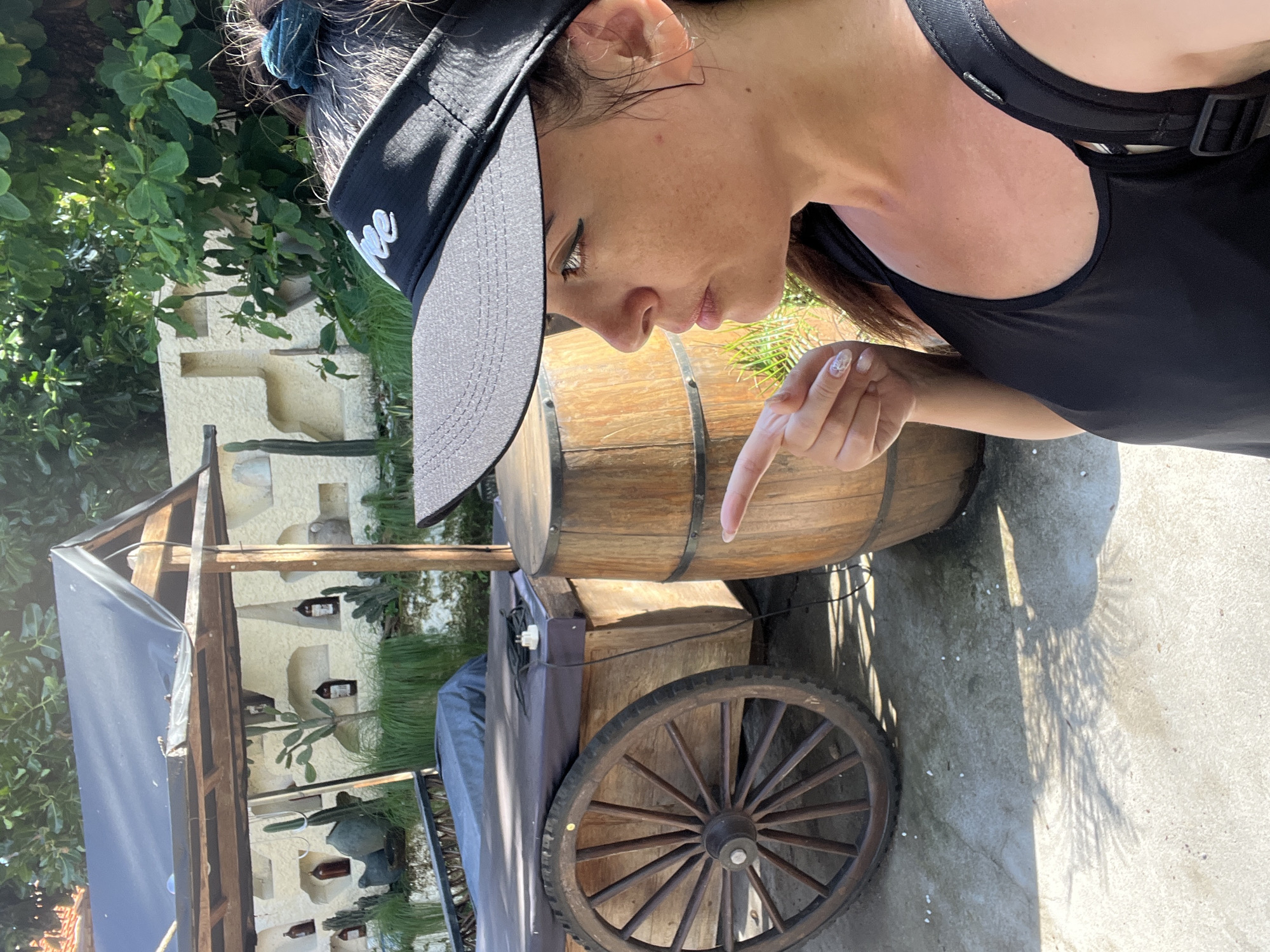 In Kuta Selatan, Indonesia, a woman is captured in a selfie, her profile visible as she points towards a rustic wooden cart and a barrel. The cart, covered with a dark tarp, features a large spoked wheel. Adjacent to it, a weathered wooden barrel stands prominently. Behind these objects, a stone wall is decorated with shelves holding various bottles and decorative cacti, suggesting a themed establishment or display. Lush green foliage forms a backdrop against the wall, with sunlight casting dappled shadows on the concrete ground. The woman, wearing a black visor and tank top, is looking away from the camera, her expression and gesture conveying a sense of observation or direction.