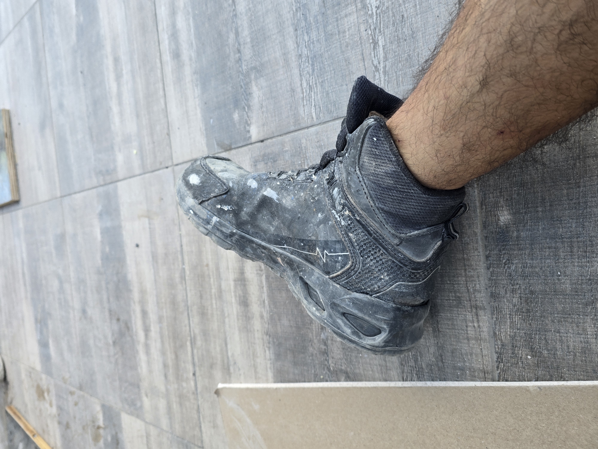The image shows a close-up, low-angle view of a person's left leg and foot, wearing a dirty black work boot. The boot is scuffed and splattered with white paint or plaster. The sock is black and pulled up to the ankle. The leg is hairy and shows a few minor scratches or marks. The boot is resting on a surface of large, grey, rectangular tiles that are laid out in a pattern. To the left of the boot, a light brown piece of material, possibly cardboard or wood, is partially visible. The background is a continuation of the tiled surface. The scene appears to be outdoors on a paved area, likely a construction site or a work area. There is no visible indication of the city of Yambol, Bulgaria.