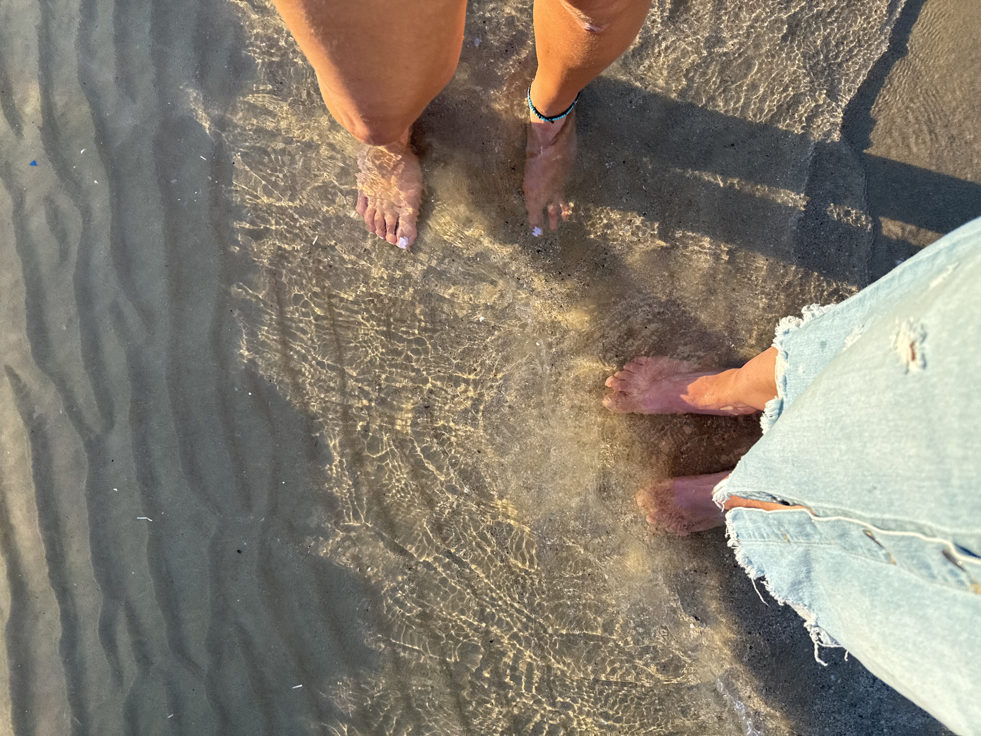 The image shows a low-angle view of two people's feet standing in shallow, clear water on a sandy seabed. The water has ripples, and sunlight refracts through it, creating patterns on the sand. One person is wearing ripped light-wash denim jeans, and the other is wearing a blue beaded bracelet on their ankle. The scene suggests a beach or a body of water with a sandy bottom, likely in Tel-Aviv, Israel, given the context.