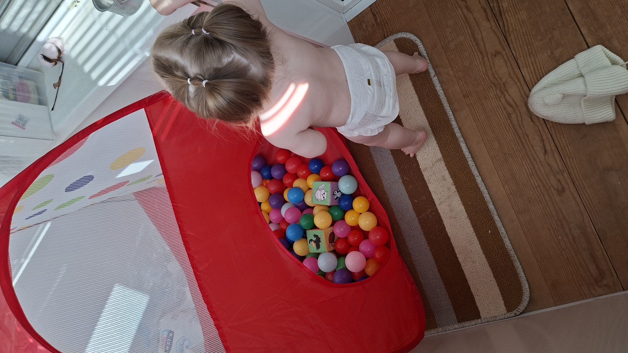 A top-down shot shows a toddler in a diaper leaning into a red, mesh-sided ball pit filled with colorful plastic balls and several blocks with animal images on them. The child has blonde hair styled in two pigtails. The scene is indoors, with a striped rug and a white knitted hat on a wooden floor. Additional items are visible near a window, including a cotton boll. The location is Chaikovsky, Russia.