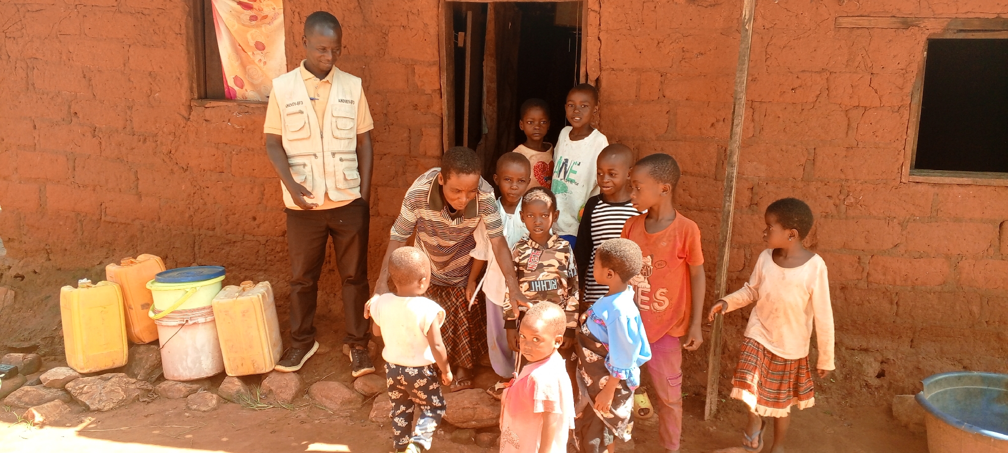 In Ngalula, Democratic Republic of the Congo, a group of people stands outside a mud-brick building. A man in a vest with the words "ANOVEV-EF3" stands to the left, while another man in a striped shirt interacts with a young child. Approximately ten children are gathered in front of the building, some looking at the camera. Yellow and white plastic containers are stacked near the building's base. The building has a doorway and a window. The ground is dirt, and there is a large tub to the right.