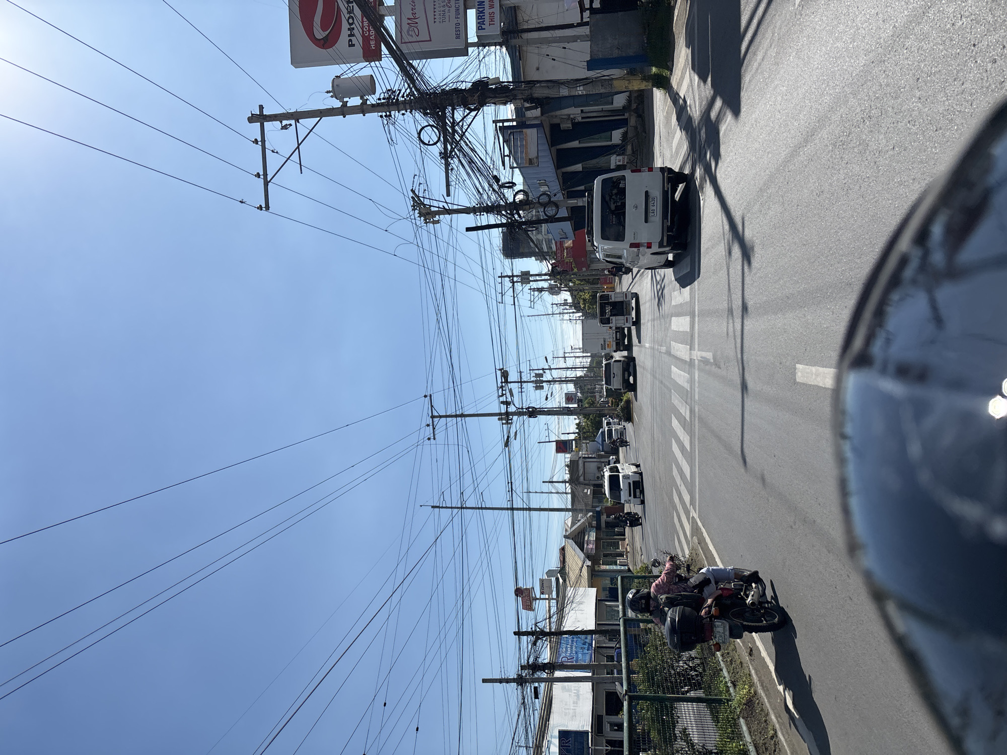 The image captures a view from a vehicle traveling on a street in Davao City, Philippines. The sky is clear and blue, with a strong presence of utility poles and a dense network of overhead wires, typical of urban areas in the Philippines. The road is paved with asphalt and marked with white lines, including a pedestrian crossing in the middle ground.
Several vehicles are visible on the road: a motorcycle with a rider wearing a helmet and a backpack is in the foreground on the left, traveling in the same direction. Further ahead, there are white vans and other vehicles. Buildings line the right side of the street, some displaying signage for businesses such as "Phoenix" and "Maria's". The architecture of the buildings suggests a commercial or mixed-use urban environment. The scene is well-lit, indicating it is daytime. The perspective suggests the photo was taken from the driver's or passenger's seat of a car.