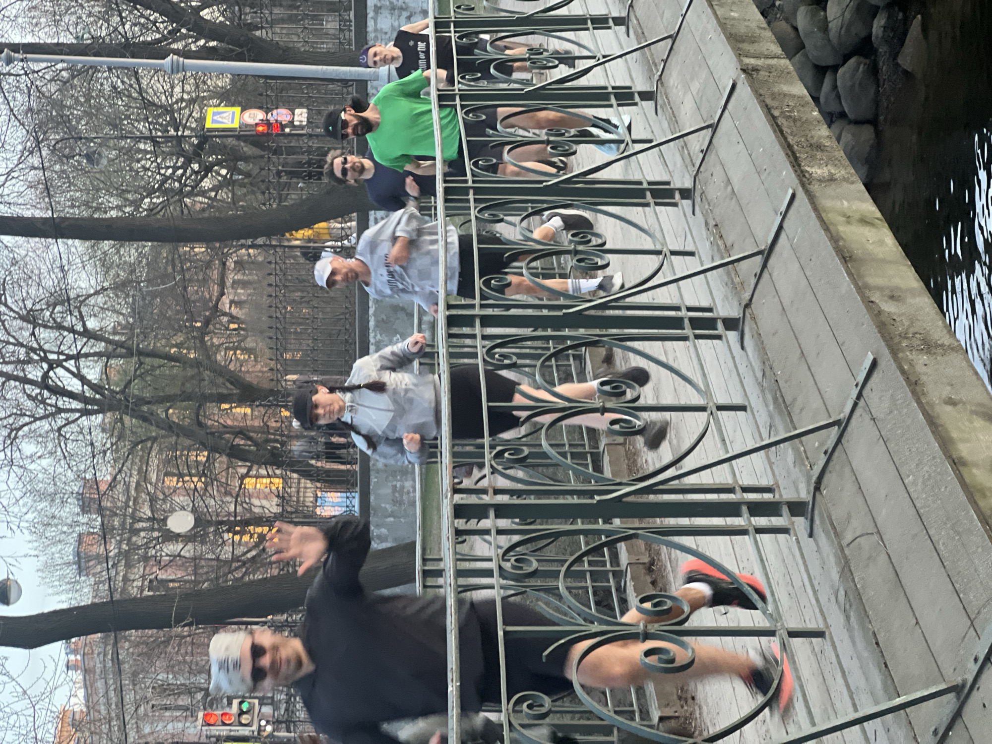 The image shows a group of six people jogging along a paved pathway in Saint Petersburg, Russia, alongside a canal or river. A decorative green metal railing separates the pathway from the water, which reflects soft light.
In the foreground, a man wearing a black long-sleeved shirt, white cap (with partially visible text "PIL"), and sunglasses is mid-stride, waving his right hand towards the viewer. Behind him, a woman with braided hair, wearing a grey and white jacket and a black cap, is also jogging. Following her is a man in a grey patterned hoodie and a white cap. Further back, a man in a bright green t-shirt, black cap, and sunglasses runs, with another man in a dark blue top and sunglasses behind him. The last runner visible is a man in a black t-shirt and a purple cap. All individuals are dressed in athletic wear.
The setting features bare trees in the background, suggesting late autumn, winter, or early spring. Beyond the trees, buildings with typical European urban architecture can be seen. Streetlights, a pedestrian crossing sign (blue square with a white figure), and red and green traffic lights are visible, indicating an active urban street environment. The light is diffused and soft, pointing to an overcast day, possibly late afternoon or early evening. The pathway is made of light-colored paving stones, and the edge of the water body is lined with large, dark stones.