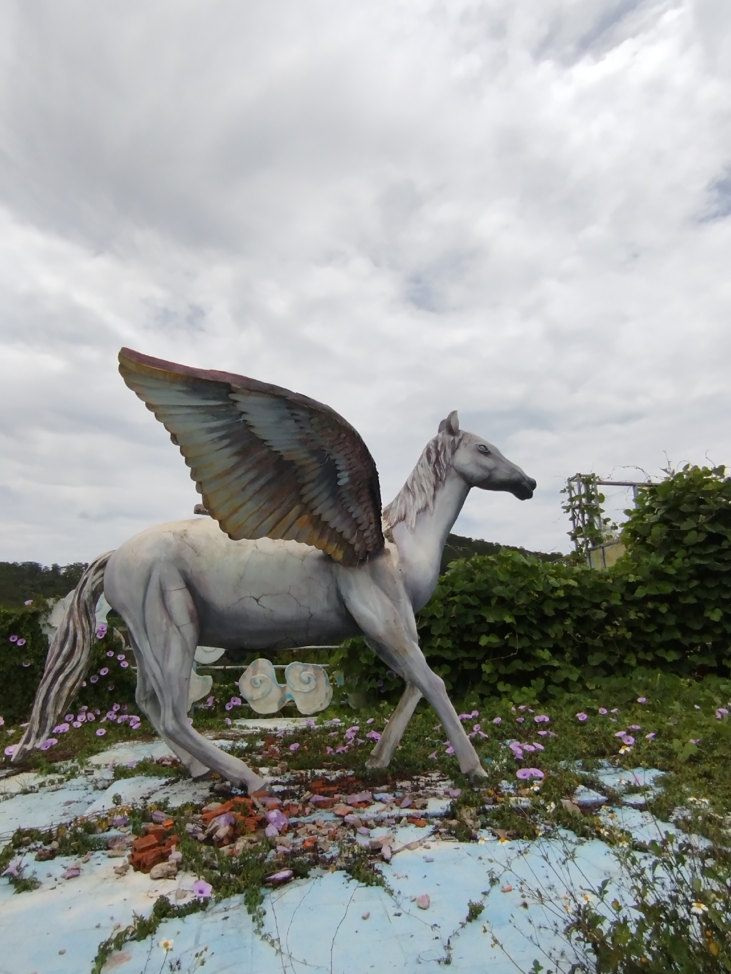 The image shows a weathered, grey statue of Pegasus, a winged horse from Greek mythology, standing on a cracked, light blue surface scattered with broken bricks, weeds, and small purple flowers. The statue has a single visible wing, tinged with brown and grey. In the background, there is a patch of dense green foliage and a rudimentary metal structure. The sky is overcast with grey clouds. The scene is located in Da Lat, Vietnam.
