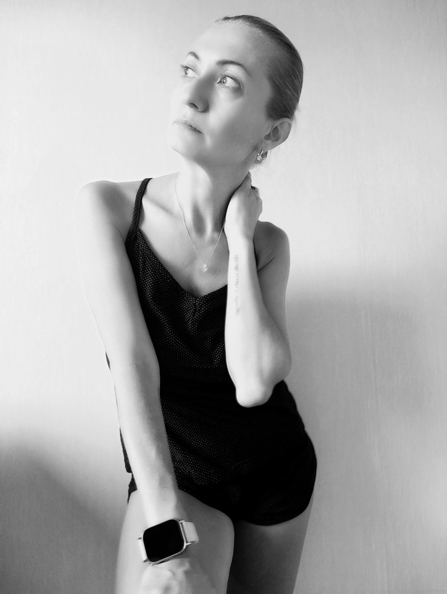 The black and white, medium shot photograph shows a woman standing in what appears to be an indoor setting, against a plain, light-colored wall. The woman is positioned in the left half of the frame, with her body angled slightly away from the camera. She has her left arm bent and held across her chest, with her left hand resting on her right arm. Her right arm is bent at the elbow, and her hand is touching her neck. She is wearing a dark, sleeveless top with a V-neckline and a delicate necklace with a small pendant. On her left wrist, she is wearing a smartwatch with a light-colored band.
The woman's head is turned upwards and to her left, with her gaze directed off-camera. Her expression appears pensive or contemplative. Her hair is pulled back into a bun. Visible on her right forearm is a tattoo of what appears to be text. She is wearing a small earring in her visible ear.
The lighting in the image is soft, creating shadows on the wall behind her. The overall mood is one of introspection. No specific time of day or weather conditions are discernible, and the setting offers no explicit clues about the location beyond being indoors. There is no discernible text in the image other than the tattoo on her arm, which is not legible. The woman's attire suggests a casual or athletic context.