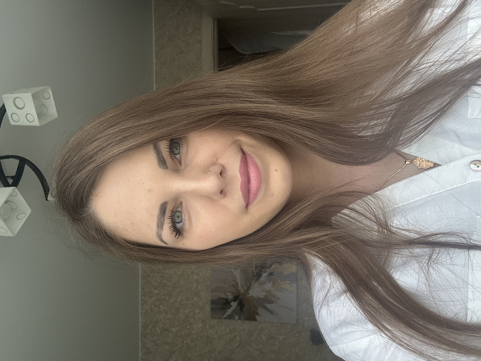 A woman with long brown hair and blue eyes is facing the camera. She is wearing a white shirt and a gold necklace. She has a nose piercing. The background includes a wall, a light fixture, a painting, and a partially visible doorway. The location is unknown and cannot be determined from the image. No information suggests that the picture was taken in Раздолье, Russia.