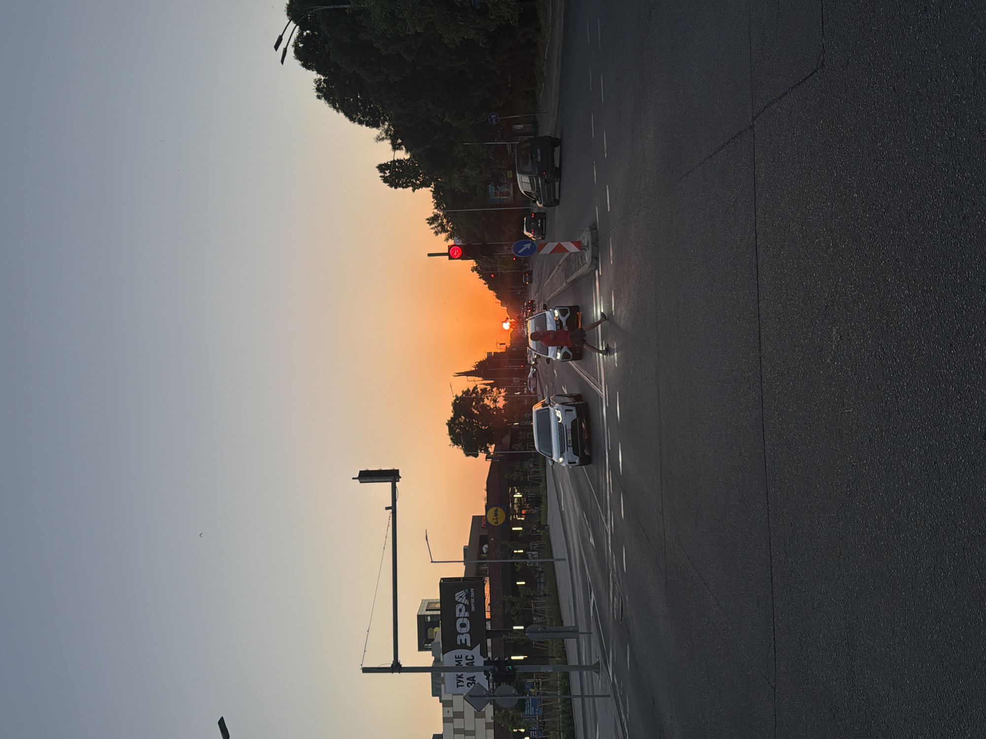 This is a photograph of a street scene at sunset in Burgas, Bulgaria. The sun is low on the horizon, casting a warm, orange glow across the sky and illuminating the road. Cars are driving on the asphalt road, and there are traffic lights and street lamps. On the left side of the road, there are trees and buildings. One building has a sign with the Cyrillic text "ЗОРА" and what appears to be a logo for a supermarket. Another sign has Cyrillic text that is partially visible, reading "ТУК МЕ ЗААС AC". The overall mood of the image is peaceful and serene, with the warm colors of the sunset creating a sense of calm.