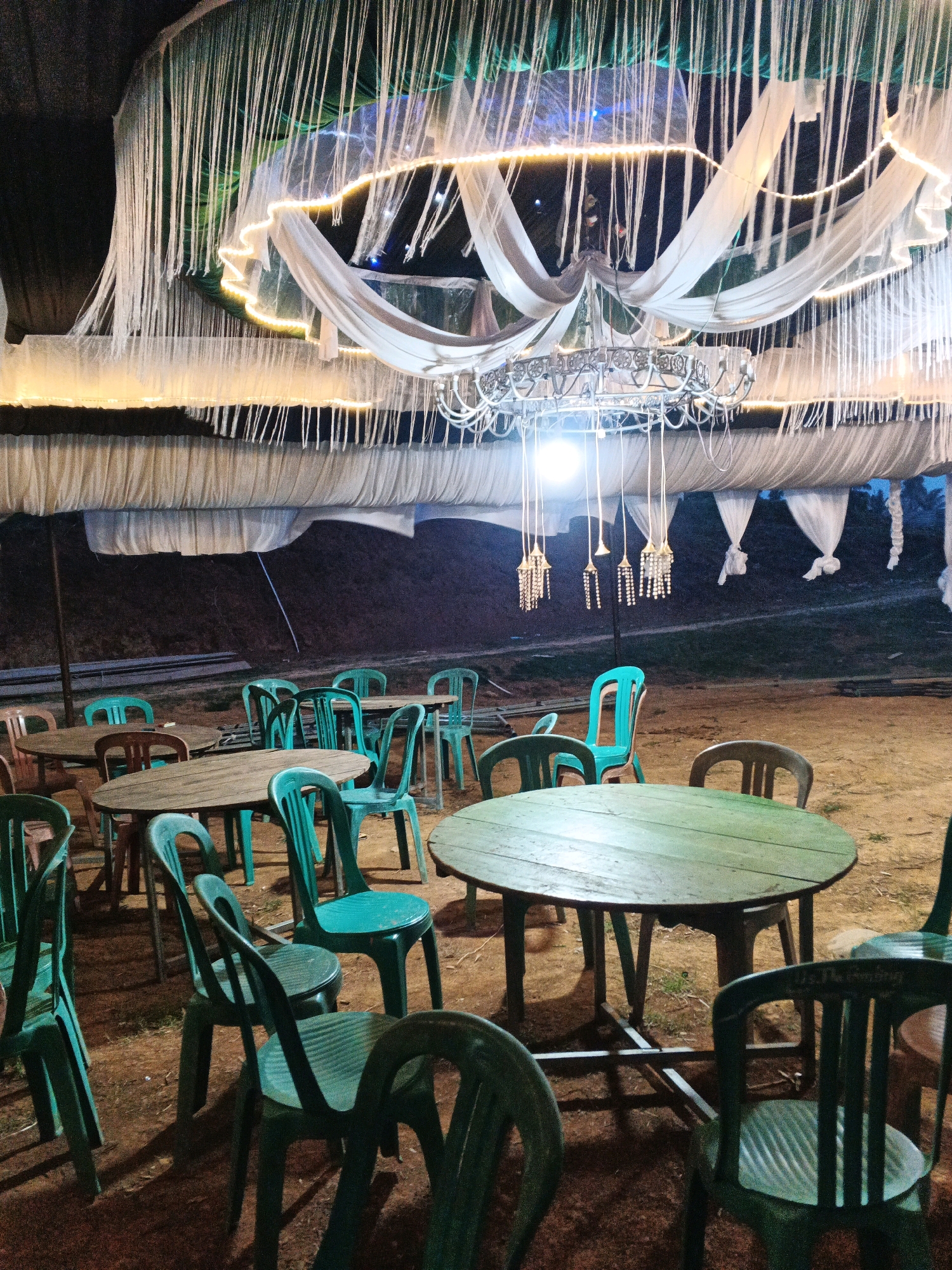 The image shows an outdoor seating area at night, likely for an event or dining, located in Brangan Mulya, Indonesia. A tent structure with white fabric draped from the ceiling is illuminated by string lights and a chandelier. Below the tent, several round wooden tables with green plastic chairs are arranged on a dirt ground. The lighting is dim, casting shadows on the ground. The background is dark and shows a slight incline of terrain. There are no people visible in the image.