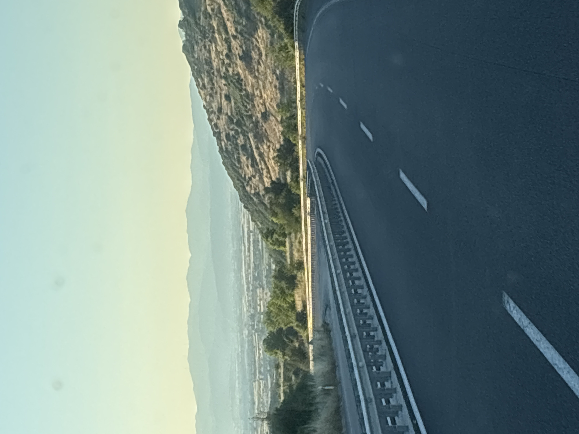 The image shows a curved asphalt road with white dashed and solid lane markings. On the left, a metal guardrail runs along the side of the road, separating it from a dry, grassy embankment. Beyond the embankment, trees and a distant valley with buildings and more hills are visible under a clear, pale blue sky. The road appears to be in a rural or semi-rural area, possibly a highway on the outskirts of the city of Avra, Greece. The lighting suggests it is either early morning or late afternoon. There are no people or vehicles in the image.