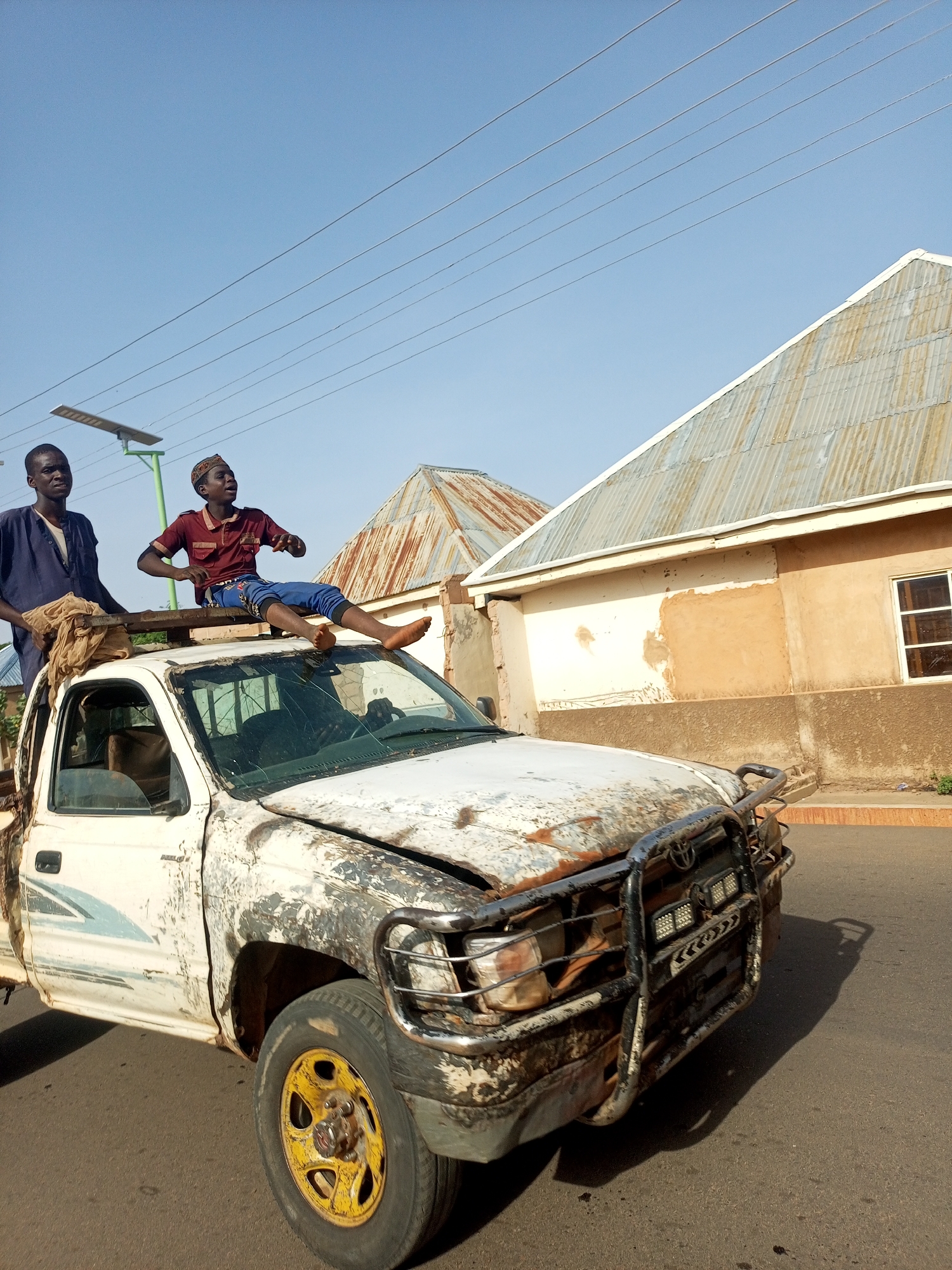 Two men are visible on and around a heavily damaged white Toyota Hilux pickup truck in a location presumed to be Tempure, Nigeria based on user prompt. The truck's paint is heavily chipped and marred, and the windshield is cracked. A man dressed in a blue robe stands on the bed of the truck. A younger man wearing a maroon shirt, blue pants, and a kufi cap sits on the truck's roof, resting his bare feet on the broken windshield. A wooden plank is positioned on the roof, serving as a seat for the younger man. In the background, a tan building with a corrugated metal roof is seen, along with power lines and a solar street light. The truck is on an asphalt road.