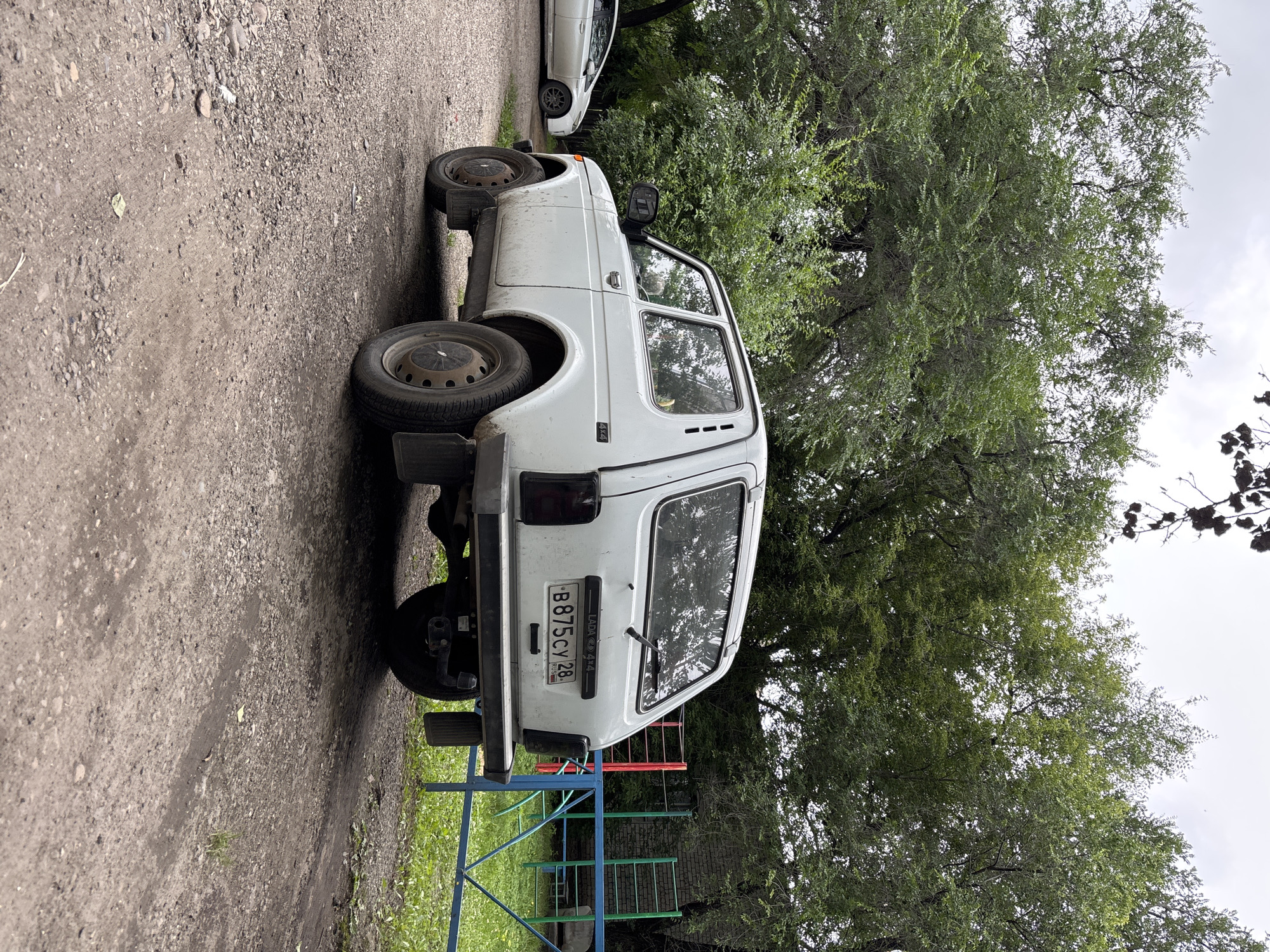 An off-white, older model Lada Niva is parked on a gravel road. The car is dirty and appears weathered. The rear of the car is visible, showing its license plate, which reads "B875CY 28 RUS". To the left of the Lada, a white modern sedan is partially visible. The background consists of lush green trees and a partially visible chain-link fence with colorful playground equipment behind it. The sky is overcast. The location is the city of Blagoveshchensk, Russia.
