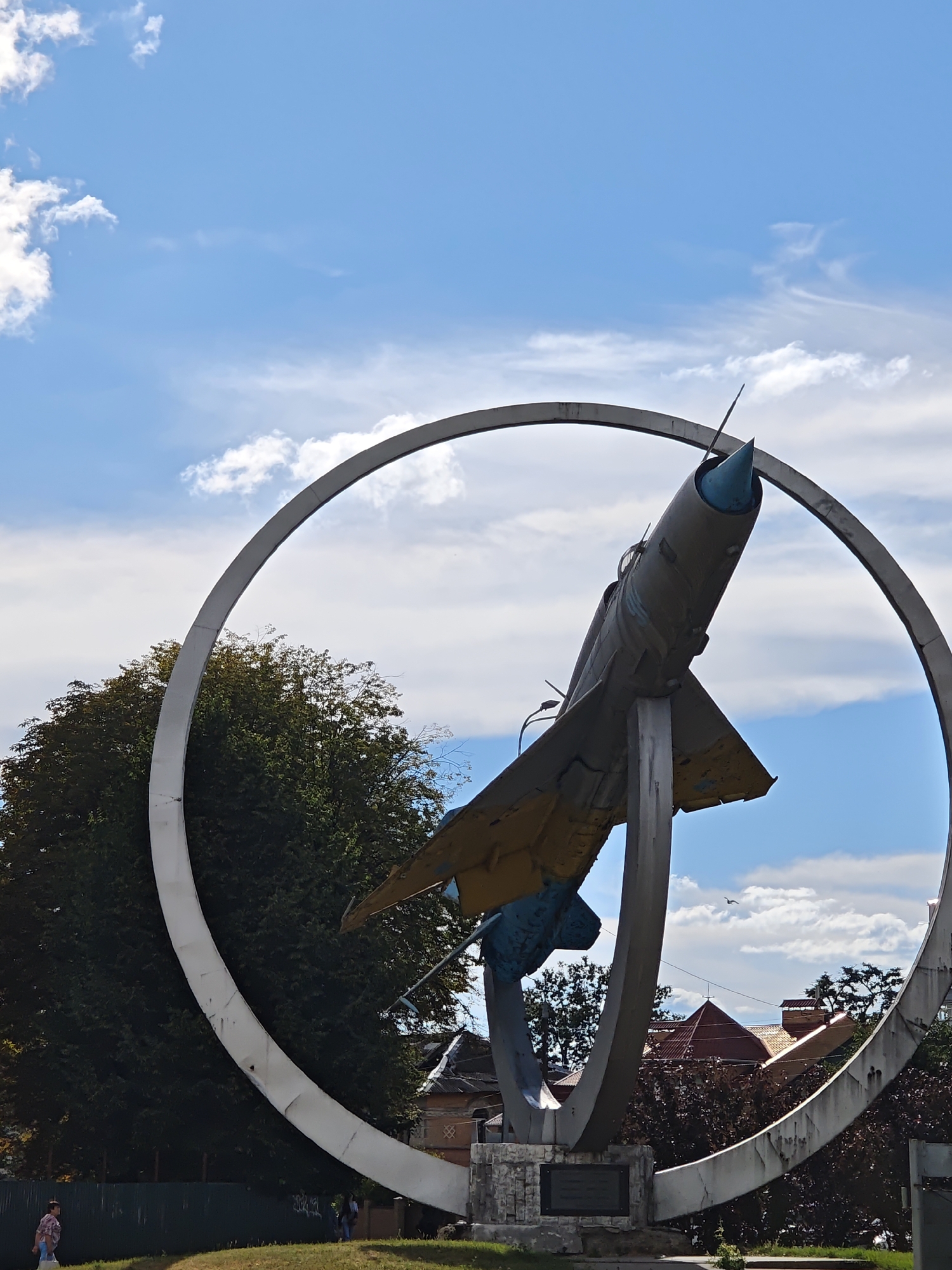 This is a low-angle, medium shot of a monument featuring a fighter jet suspended within a large, circular frame, set against a backdrop of a bright blue sky with scattered white clouds. The jet, painted primarily in faded olive green and yellow, is angled upwards and to the right, with its nose cone a bright blue. It is supported by a concrete pedestal. The circular frame, made of a light-colored material, is incomplete, appearing as a broken arc that extends from the lower left to the upper right of the frame. In the background, a dense green tree is visible behind the left side of the circular frame, and behind the jet and pedestal, there are several buildings with red-tiled roofs, suggesting an urban or suburban setting. The sky is clear except for a few wispy clouds and a distant bird in flight. In the foreground, a patch of green grass leads to a dark green fence. To the far left, a person in a light-colored plaid shirt walks away from the camera, and further to the right, two individuals stand near the fence. The monument is located in Vinnytsia, Ukraine, specifically at the intersection of Soborna Street and Halytskoho Square, commonly known as "Aviators Square." It commemorates the pilots of the 48th Tactical Aviation Brigade.