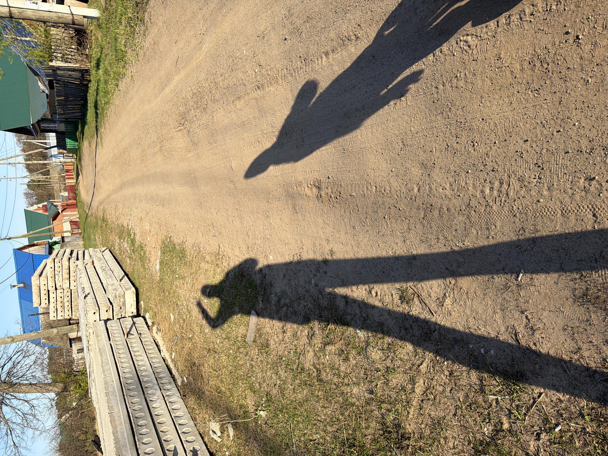 The image depicts a dirt road in what appears to be a residential area, possibly in the city of Chaikovsky, Russia. The road is flanked on one side by a stack of concrete slabs with circular indentations, and on the other side by houses, some with blue and green roofing. The sun is shining, casting shadows. In the foreground, there are two distinct shadows: one that is closer to the camera, appearing to be of a person holding a phone or object; and a second, smaller shadow further down the road, more difficult to interpret. The road shows signs of vehicle traffic. Some grass and small vegetation are present along the edges of the road. The sky is visible as blue in the background.