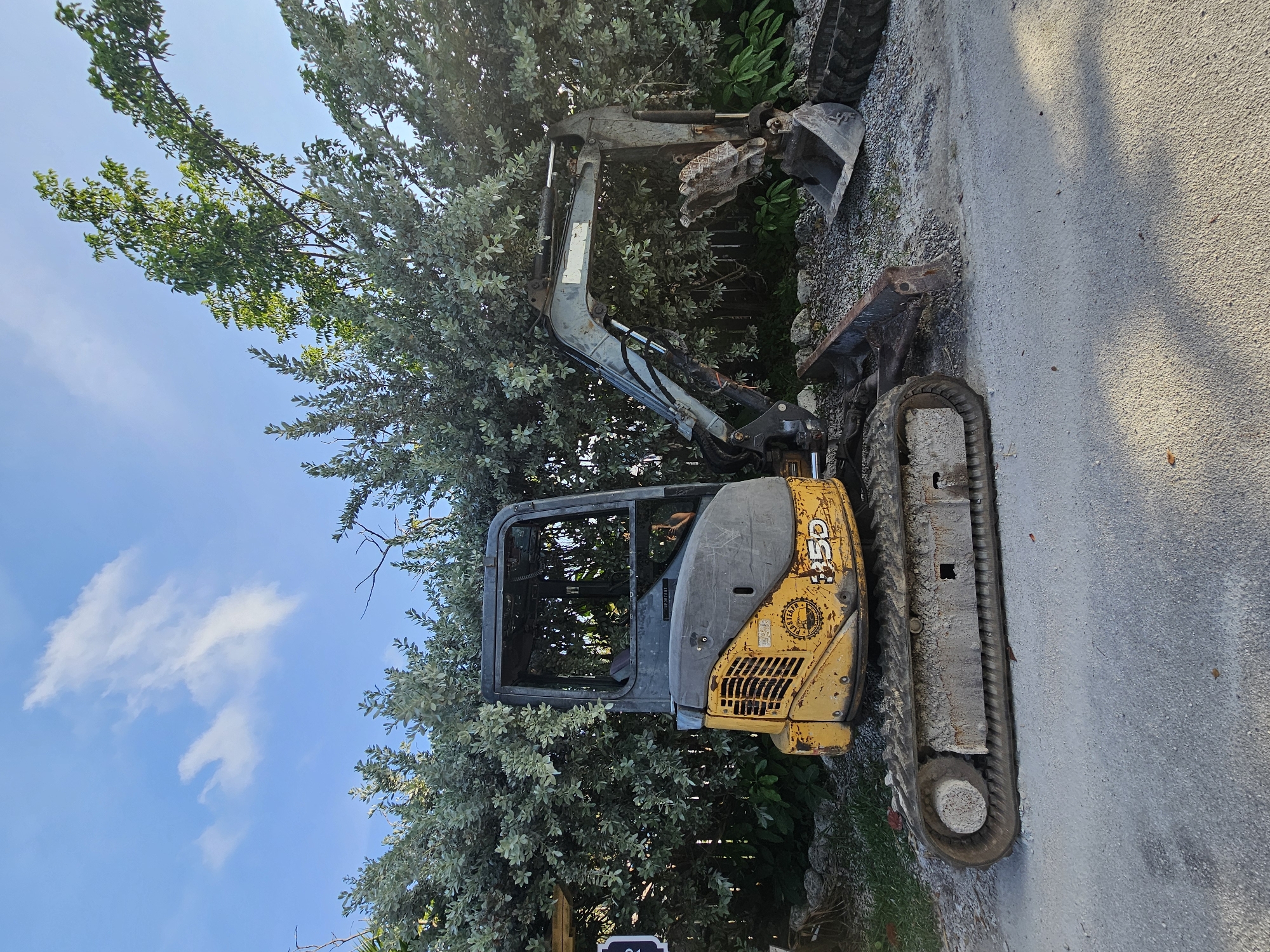 A yellow and gray mini excavator is parked on a gravel and asphalt surface in front of a dense green hedge under a blue sky with scattered clouds. The excavator is positioned at an angle to the right, with its tracks on the ground. The cab of the excavator is open at the top, and its boom is extended. A bucket is attached to the boom, and another bucket is visible on the ground to the right of the excavator. A portion of a tire is also on the ground to the right. The overall scene suggests an outdoor setting, possibly a construction site or a storage area, in Islamorada, United States of America.