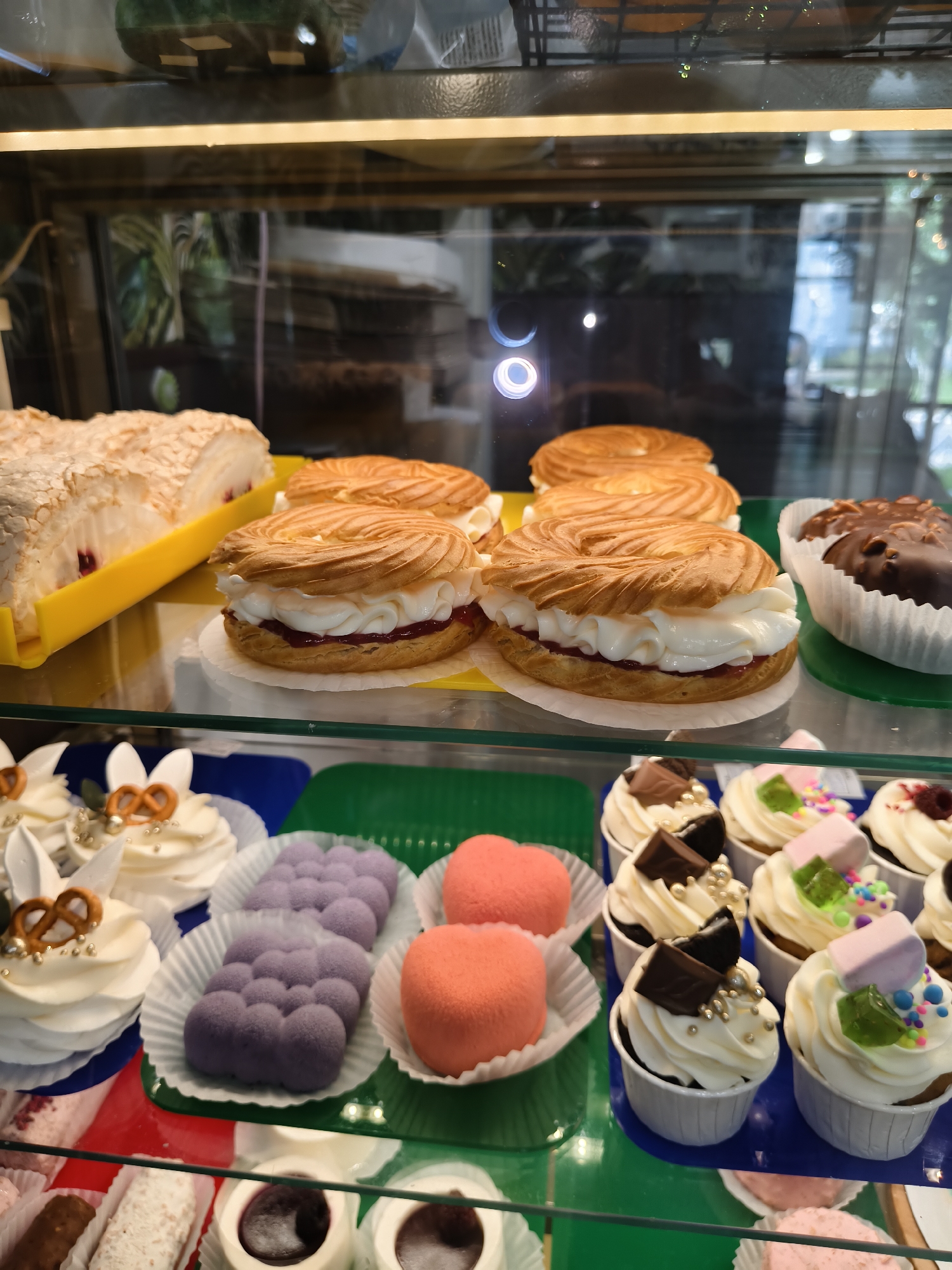 The image shows a display case filled with various pastries. In the foreground, on a green shelf, are several white cupcakes topped with cream, sprinkles, and chocolate pieces. To the left of the cupcakes, on a blue shelf, are purple, bubble-shaped pastries and pink heart-shaped pastries. Behind these, on a yellow shelf, are several ring-shaped pastries filled with cream and jam, and a slice of meringue. On the far right, there is a chocolate-covered pastry. The background is blurred, showing reflections of plants and lights.