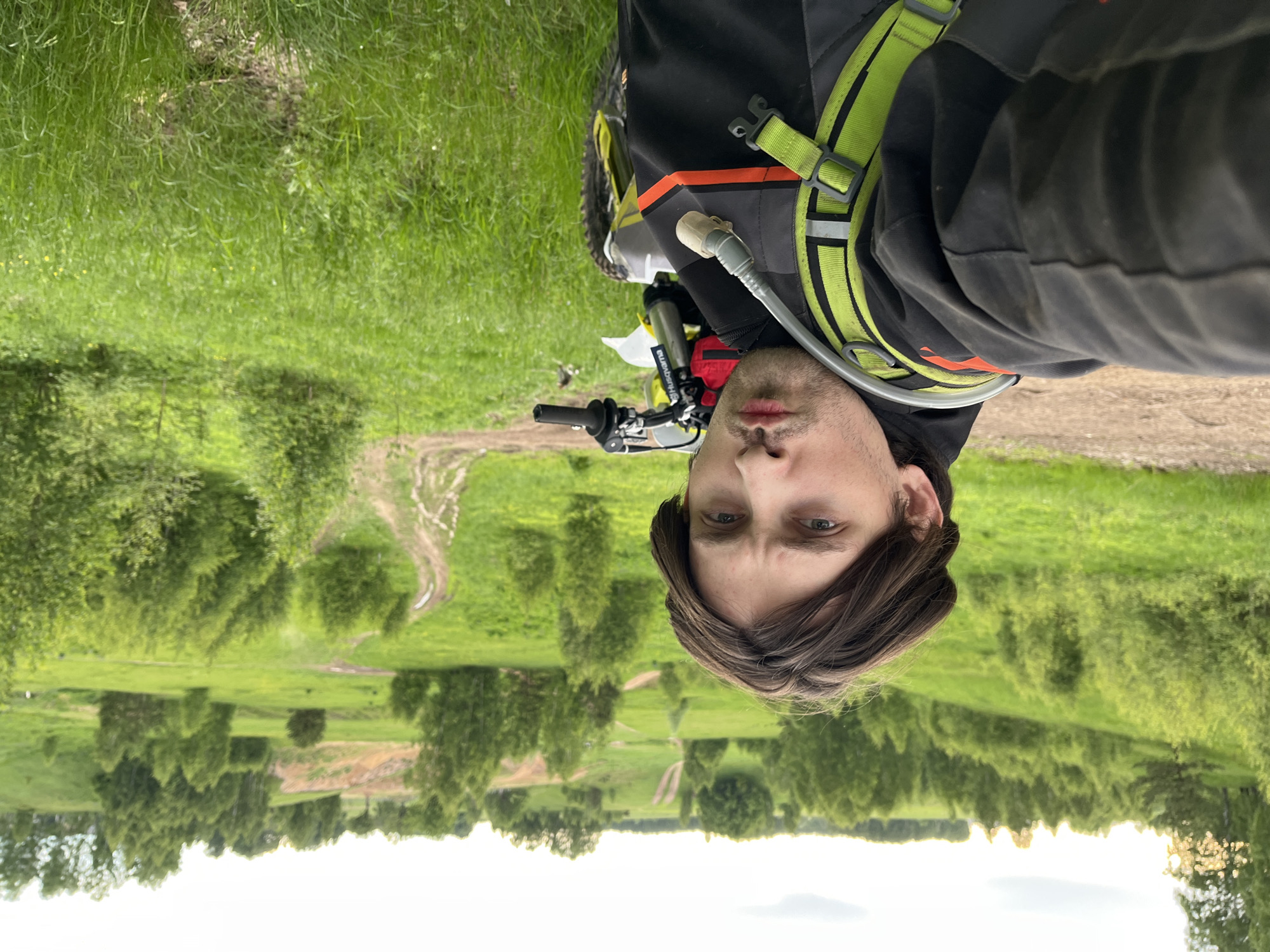 A close-up, portrait-style photo shows a man on a motorcycle in Новочувашский, Russia. The image is rotated 180 degrees. The man has brown hair and light eyes. He is wearing a black jacket with orange and grey accents, along with a lime green harness. The front of his motorcycle is visible, including the handlebars and front wheel. The background consists of a grassy field with trees and a dirt path. In the distance is a treeline against a light sky.