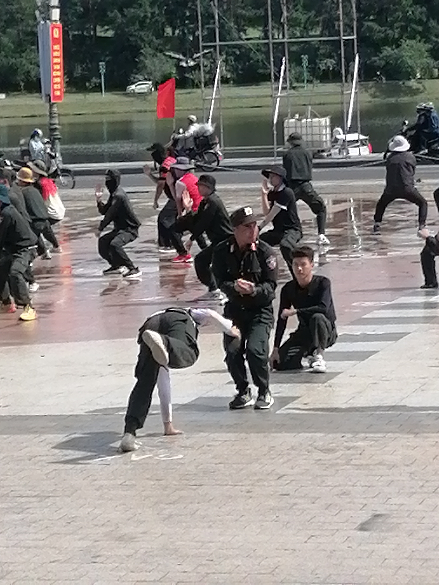 In Da Lat, Vietnam, a group of people are engaged in a coordinated activity on a wet, paved plaza, likely a public square or walkway. The ground is reflective, suggesting recent rain or water spillage. The individuals are dressed in dark clothing, with some wearing what appear to be uniforms or tactical gear, including hats and dark pants. Several individuals are in motion, performing various poses and movements, some appearing to be part of a dance or martial arts demonstration.
In the foreground, a person wearing dark pants and a light-colored top is bent over, performing a move that involves balancing on one leg. To their right, another individual in dark clothing is crouched low, facing away from the camera. Further back, several other figures are visible, some in mid-movement, while others stand or crouch. One person in the middle ground is wearing a dark uniform with a badge on their hat and appears to be observing the activity.
The background reveals a body of water, possibly a lake or canal, bordered by trees and greenery. Across the water, there are some structures, including scaffolding and what looks like a white tank or container. A red flag is visible on a pole in the distance. The overall scene suggests an outdoor public event or performance taking place in Da Lat.