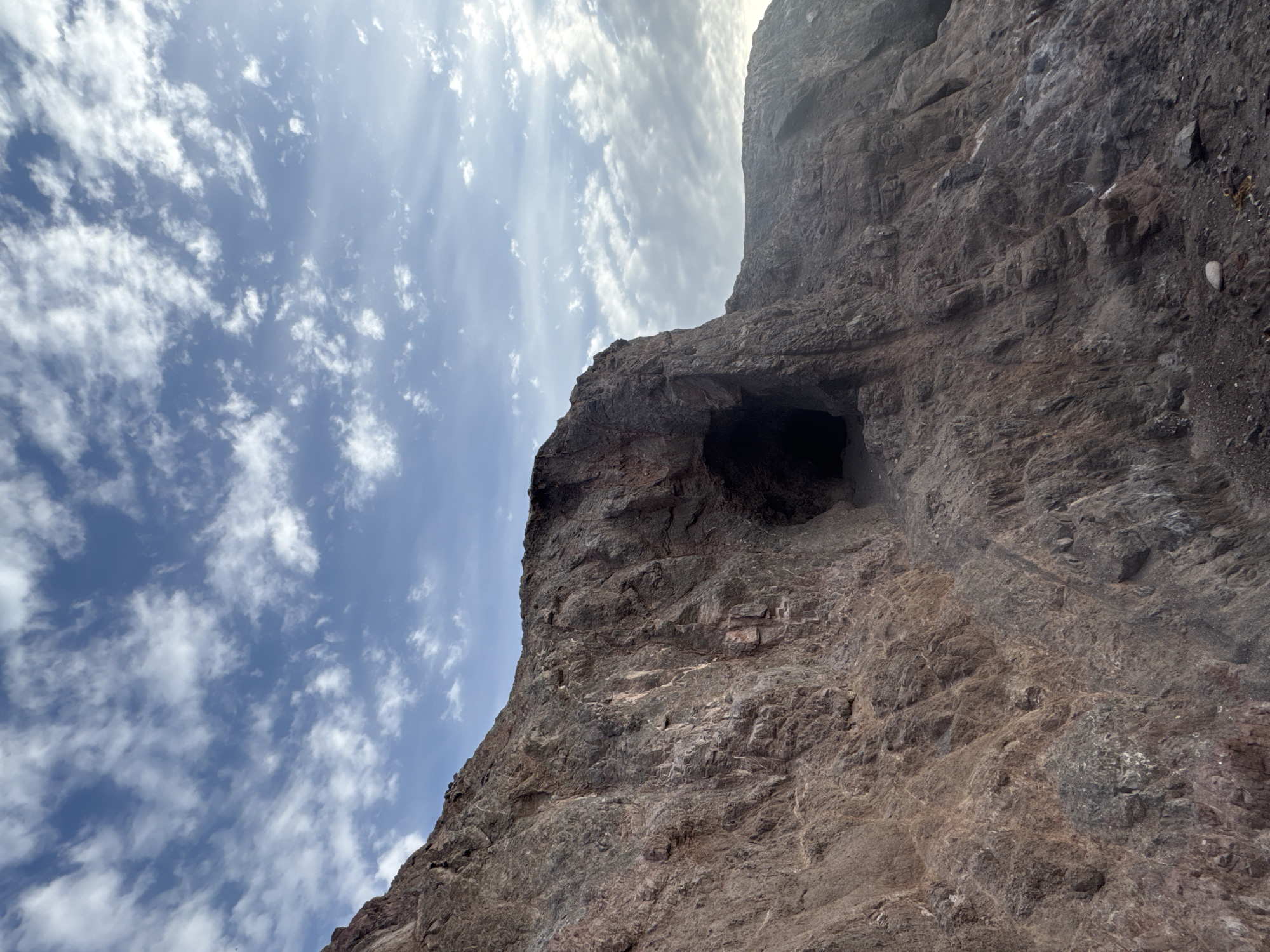 The image shows a rocky cliff face under a sky with scattered clouds. The cliff is predominantly grey and brown, with a visible cave opening. The sky is blue with white clouds scattered throughout. The image appears to have been taken from a low angle. The location and city are unknown.