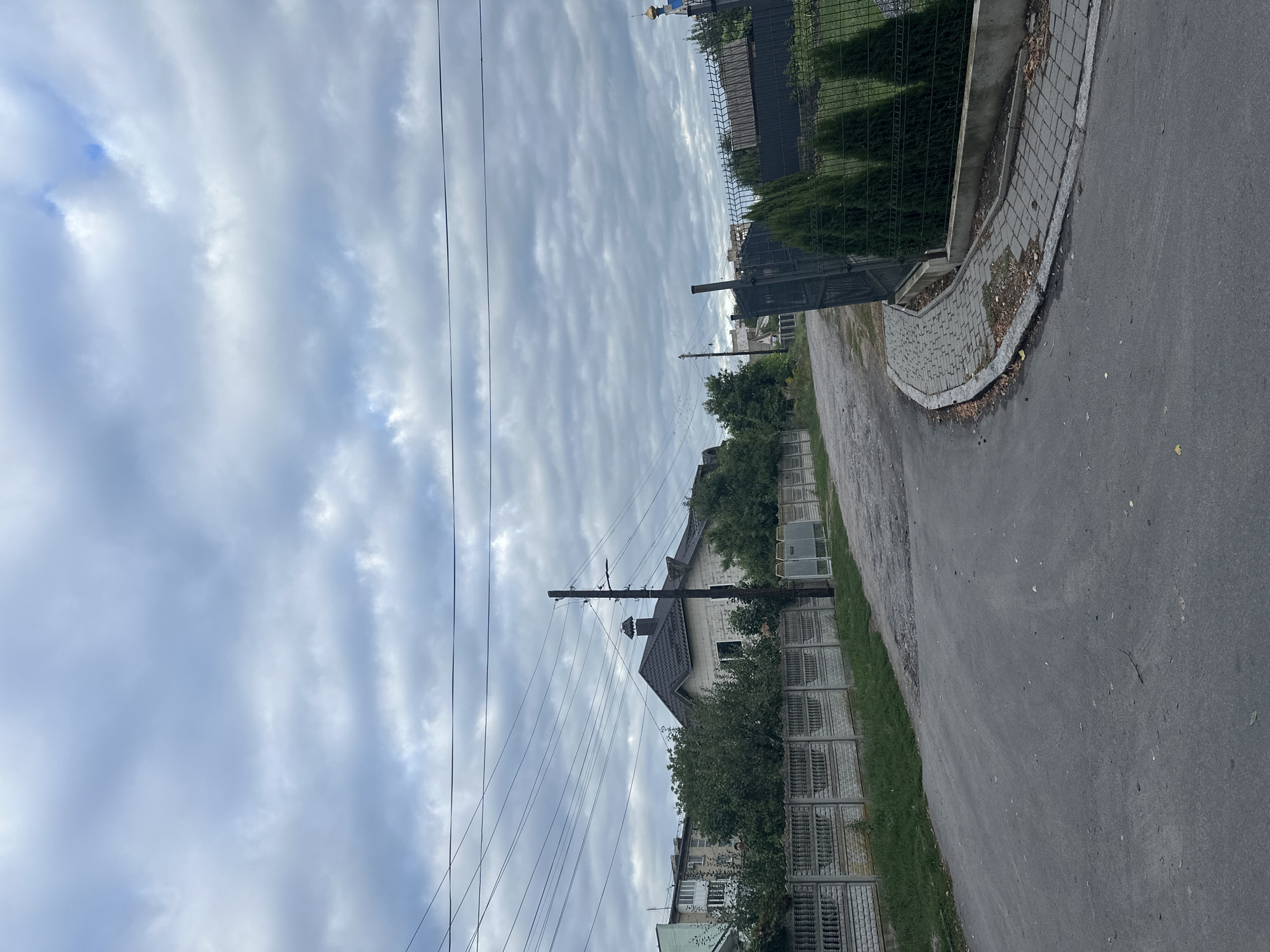 The image depicts a street scene in Khmilnyk, Ukraine, under a cloudy sky. A paved road curves to the right, bordered by a concrete fence on the left and a black metal fence with decorative cypress trees on the right. Beyond the concrete fence, a residential building with a grey tiled roof and brick walls is visible, surrounded by green trees. Further in the background, another taller residential building can be seen. A utility pole with electrical wires stands near the center of the image, positioned between the road and the residential structures. The ground along the roadside is covered in grass and scattered leaves. The scene appears to be a quiet suburban or semi-urban area.