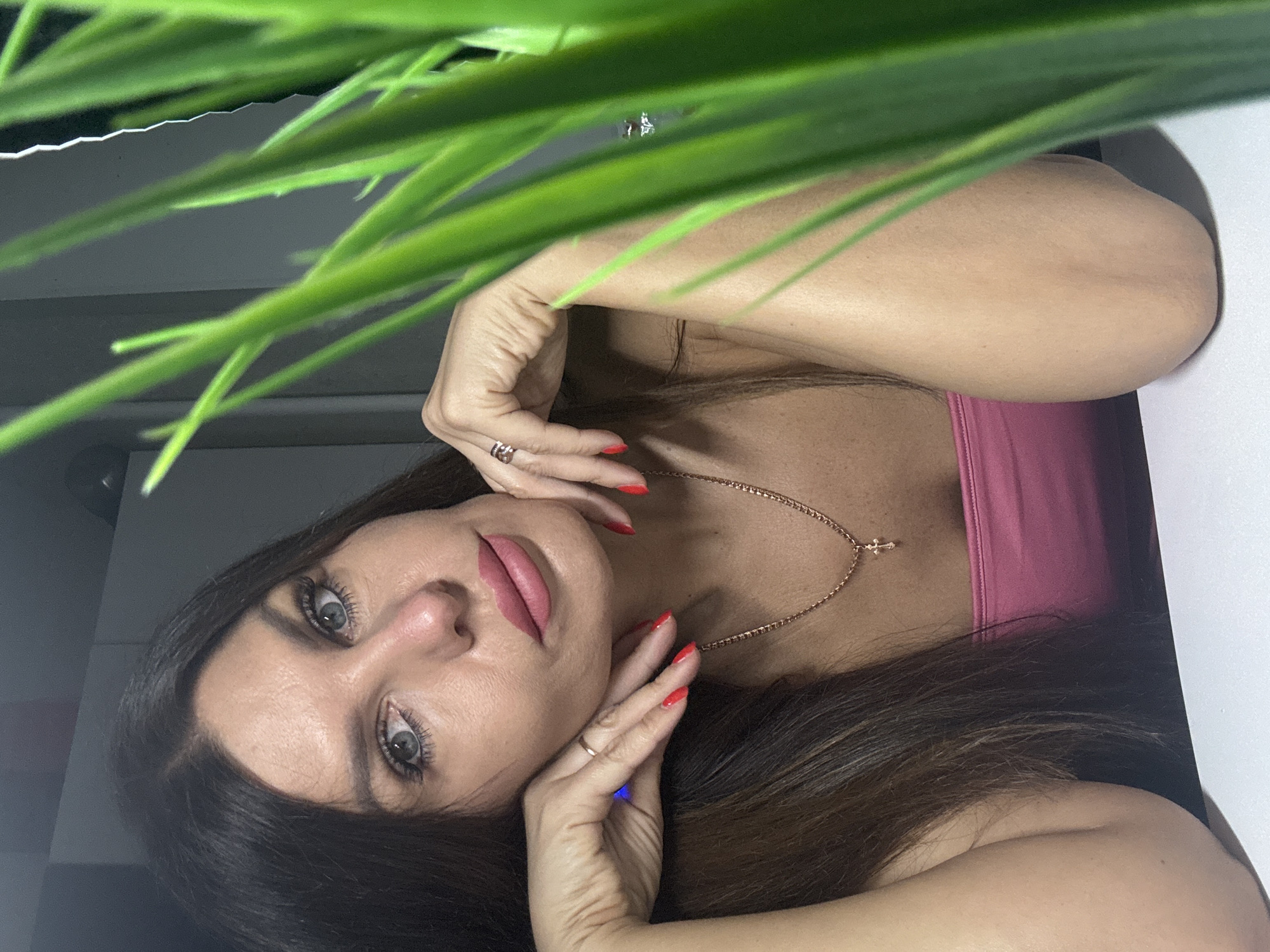 A woman with dark hair and light blue eyes is looking at the camera with her chin resting on her hands. She is wearing a pink top and a gold necklace with a cross pendant. A green plant is in the foreground, partially obscuring her face. The background is dark and out of focus. There is no indication of the location within the city of Chaikovsky, Russia.
