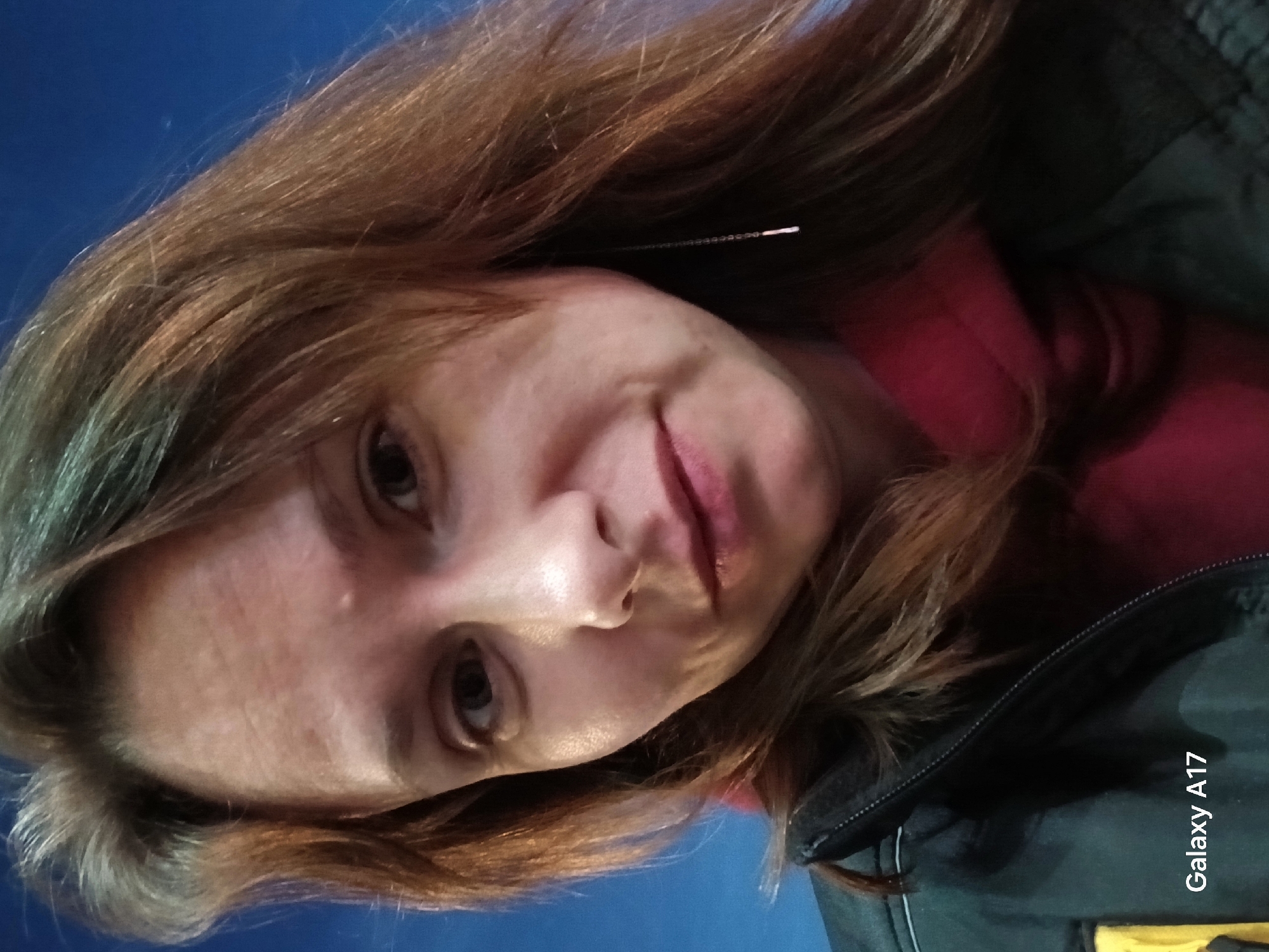 A woman is facing the camera, angled downwards slightly to the left. She has light skin, brown hair, and is wearing a dark jacket over a red top. A single straight earring is visible. The background is a solid, dark blue. "Galaxy A17" is printed on the lower right of the image. Location: Unlikely Yekaterinburg, Russia, as the camera model, Galaxy A17, is printed on the image.