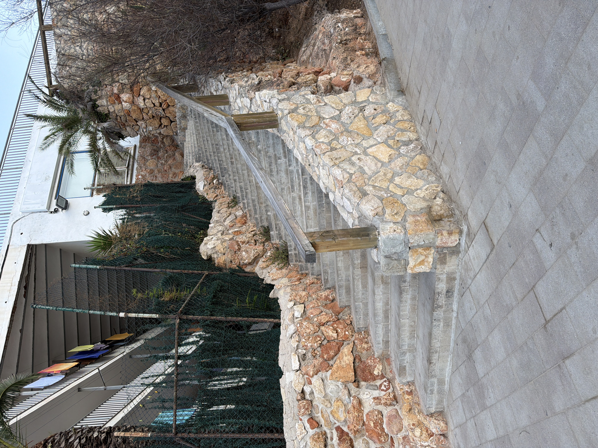 The image depicts an outdoor stone staircase with a wooden railing, leading up towards a building. The staircase is flanked by a stone wall on the left, constructed with rough, irregular stones of varying colors, predominantly orange and brown. Green foliage and a chain-link fence are visible behind the stone wall, partially obscuring the lower levels of the building.
The building itself is white with a white railing along its balconies, where several colorful garments (blue, yellow, orange, and black) are hanging out to dry. A palm tree with dark green fronds grows from a cluster of stones on the right side of the staircase. Further to the right, bare branches of a shrub or tree are visible against a rocky slope. The ground in front of the staircase is paved with rectangular grey tiles.
The scene appears to be in a residential or resort area, given the building's design and the presence of laundry drying. Salou, Spain, is a coastal resort town known for its beaches and tourist accommodations, which aligns with the visual elements in the image. The lighting suggests it is daytime, with the sun casting soft shadows. There are no people visible in the image. The overall impression is one of a functional, outdoor space, likely part of a holiday complex or residential building.