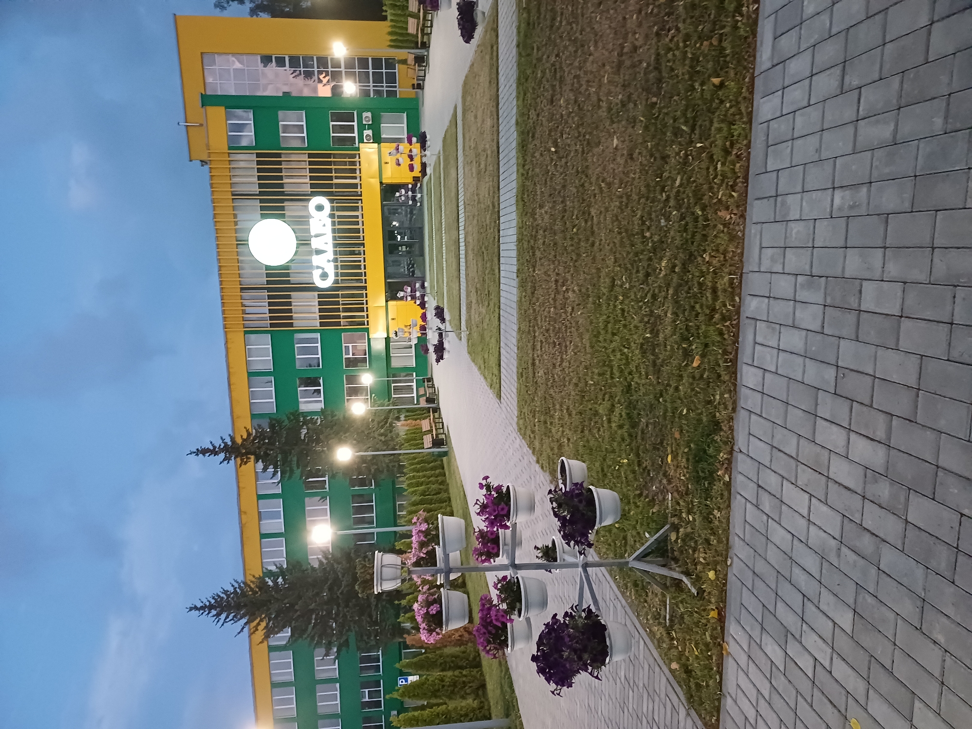 The image shows the exterior of a building in Kobryn, Belarus, with the name "СЛАВО" illuminated above the entrance. The building is painted green and yellow, with multiple windows. The sky is overcast and appears to be dusk. In front of the building, there is a paved walkway, a lawn, and several planters filled with purple flowers. There are also lampposts and benches visible in the scene. The overall impression is of a well-maintained, modern building with a landscaped outdoor area.