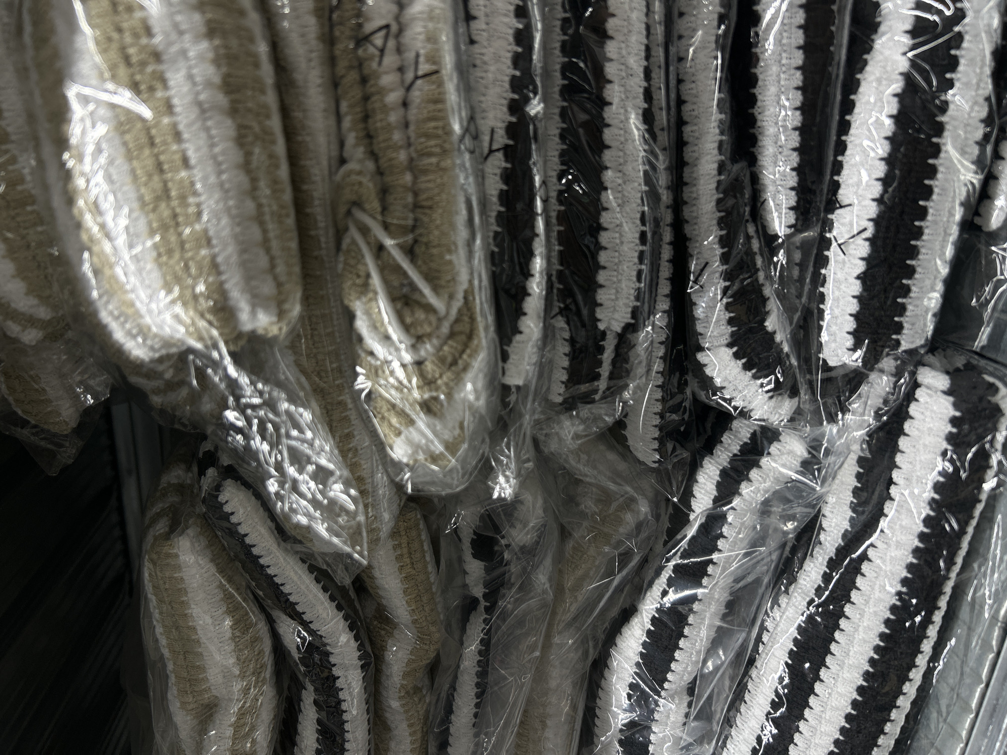 The image displays a close-up view of stacked, individually plastic-wrapped fabric items, likely towels or blankets, on shelves. The items feature horizontal stripes in alternating colors of black and white, or beige and white. The shelves are metal and appear to be in a storage or retail environment. The lighting is dim and direct, creating some glare on the plastic wrapping and highlighting the textures of the fabric. There are no people or specific identifiable landmarks present, therefore the location within Holon, Israel cannot be determined from the image alone.